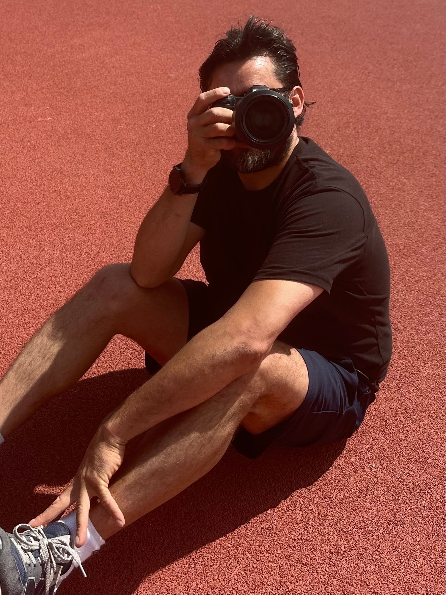 Male photographer sitting on red track taking photo with DSLR camera wearing black shirt and blue shorts.