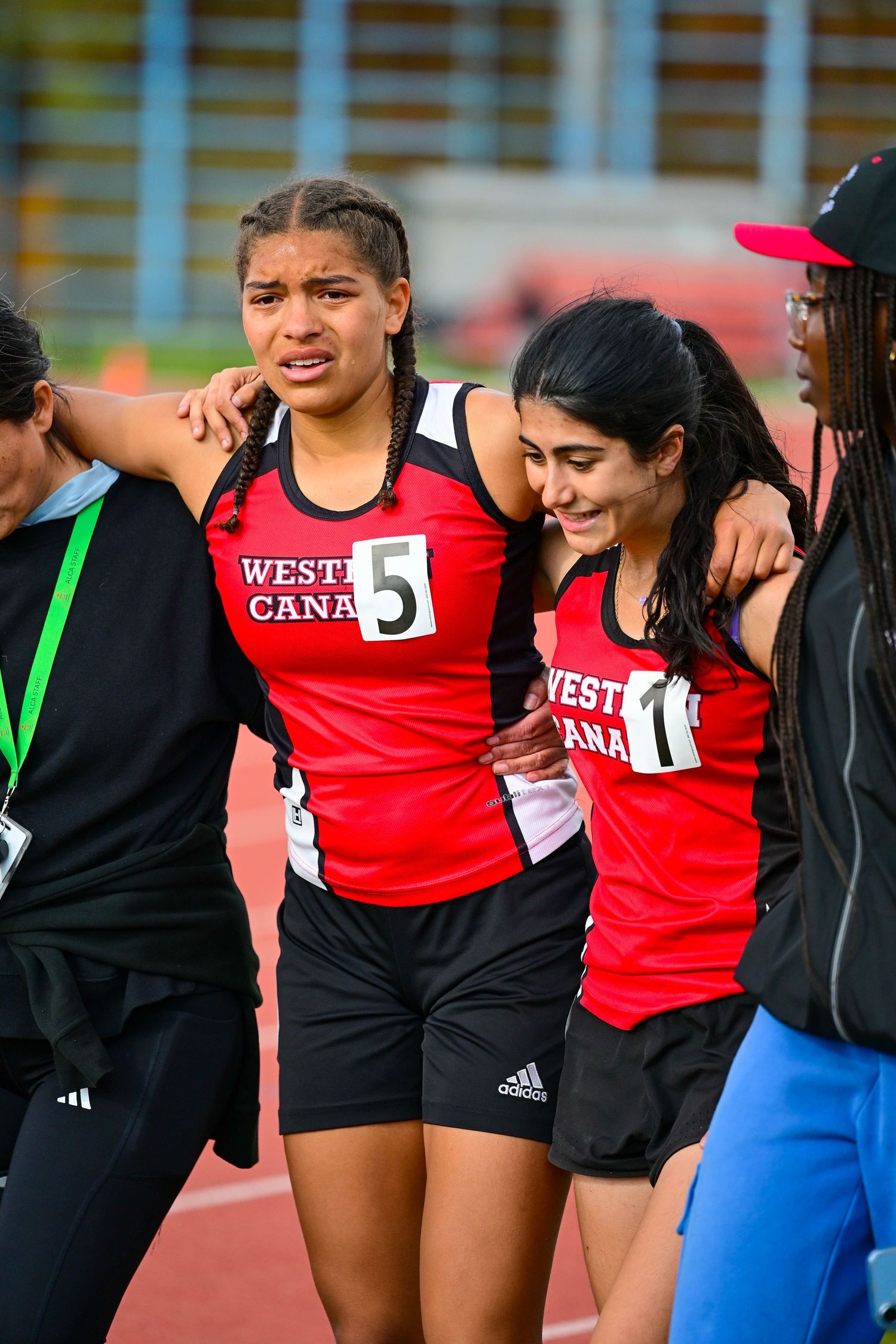 Two Western Canada female track athletes in red uniforms embracing emotionally after competition on track.