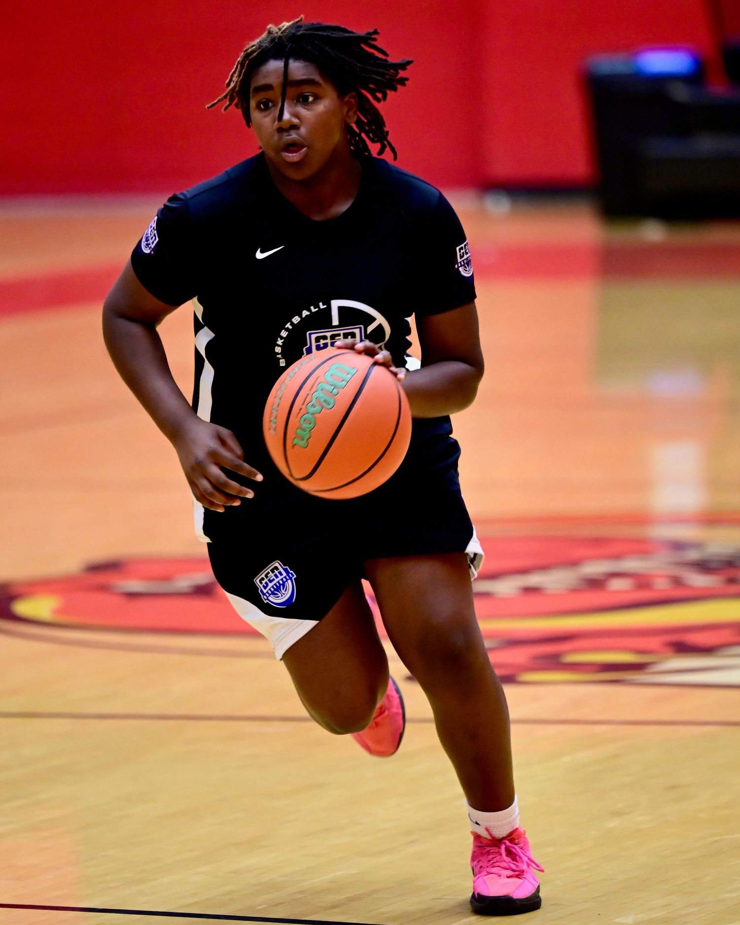 A young female basketball player dribbles a ball during a practice session in a red gymnasium.