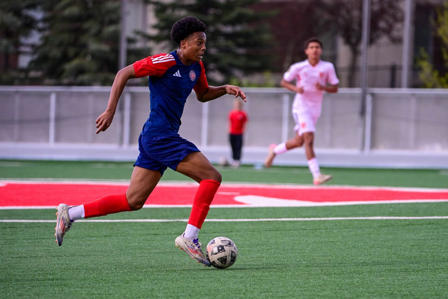 Young soccer player in blue and red uniform dribbles ball on red turf field during match.