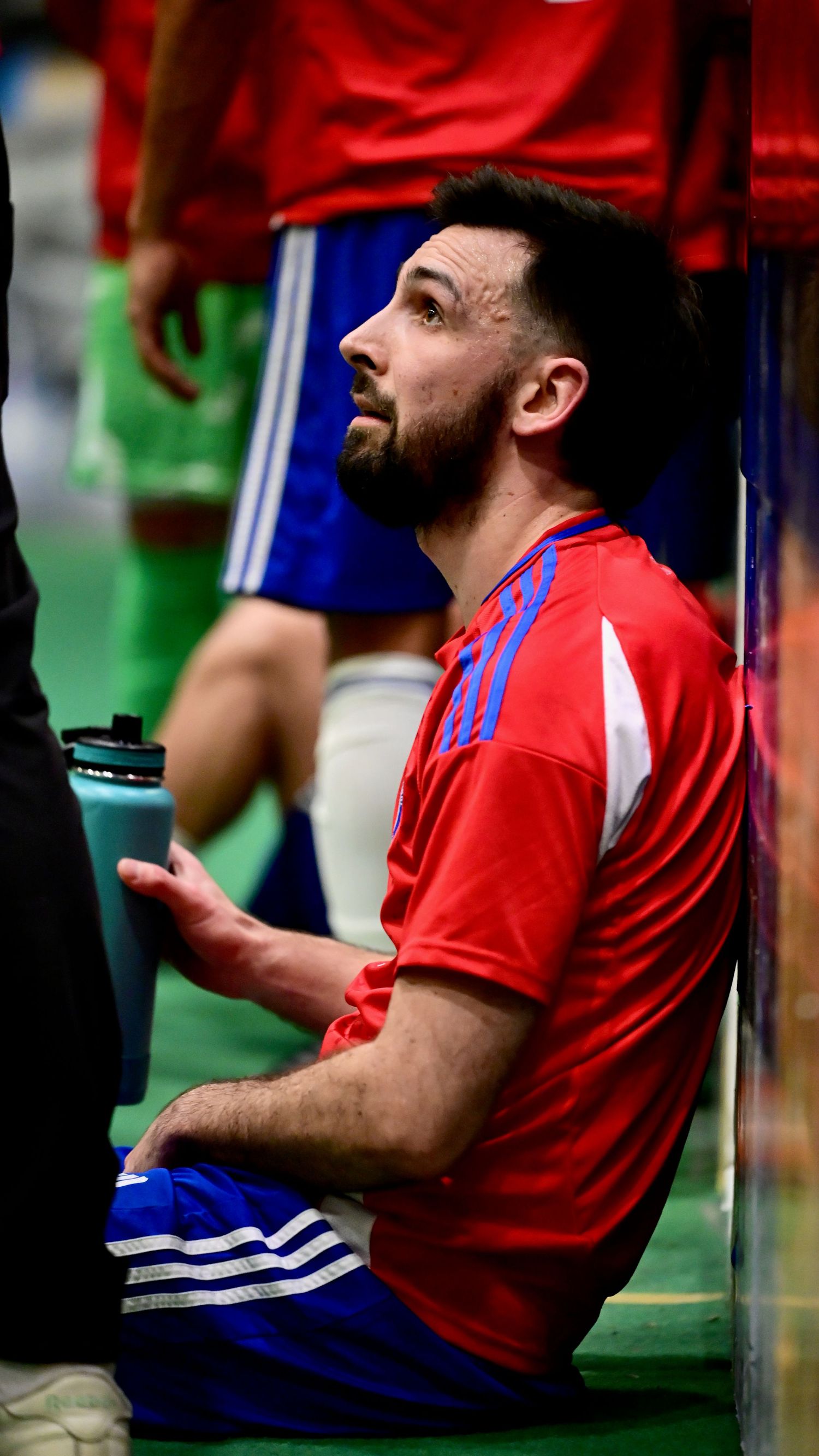 Male soccer player in red Adidas jersey sitting against wall looking up during indoor futsal game.