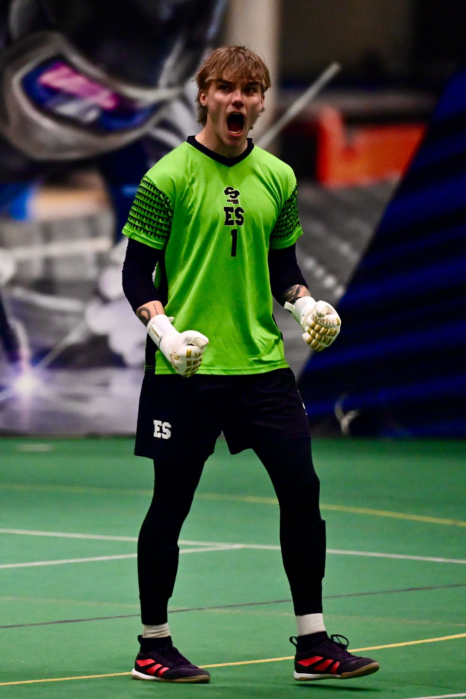 Indoor soccer goalkeeper in bright green jersey celebrating with mouth open and fists raised.