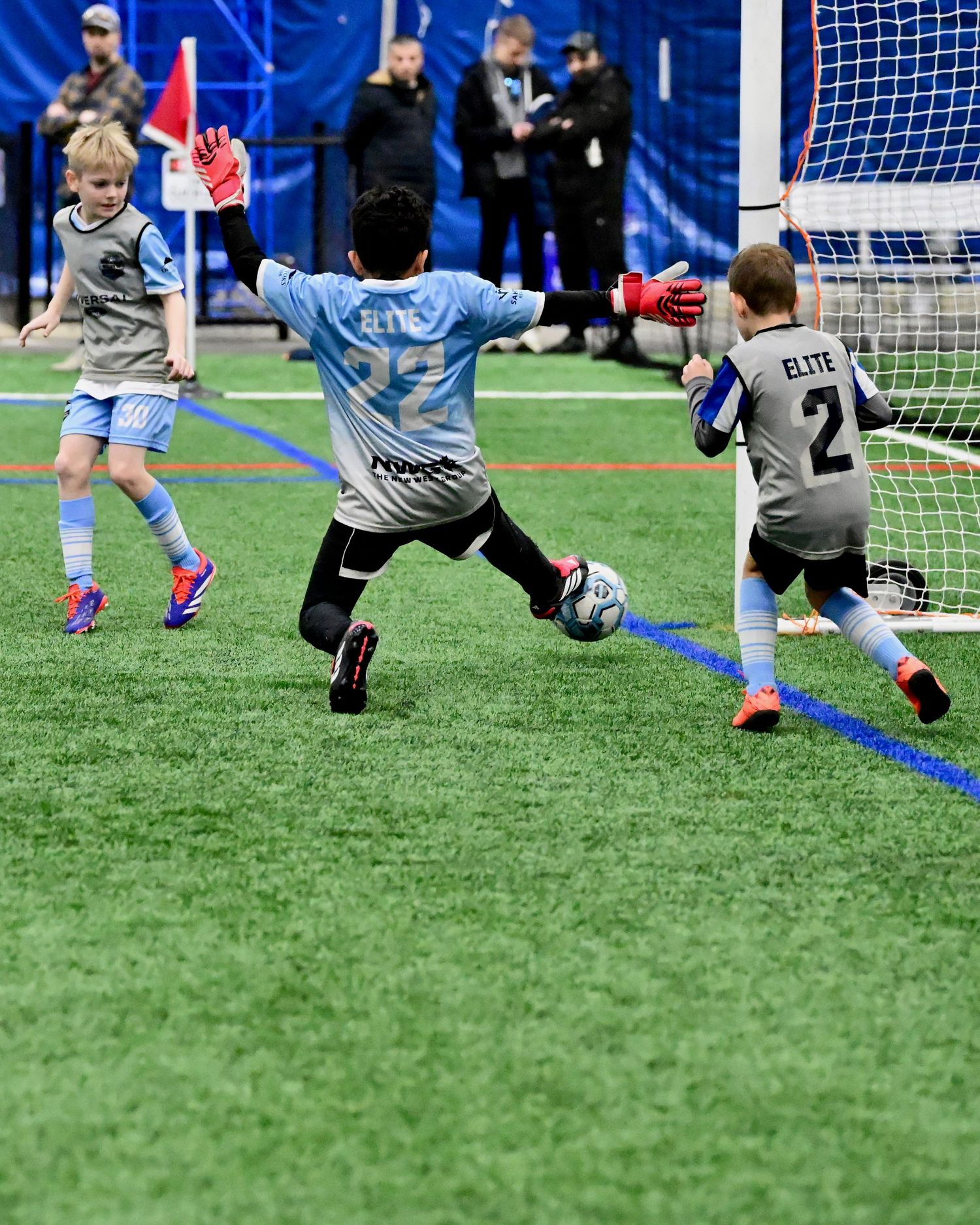 Youth indoor soccer goalkeeper blocks shot during competitive match on turf field.