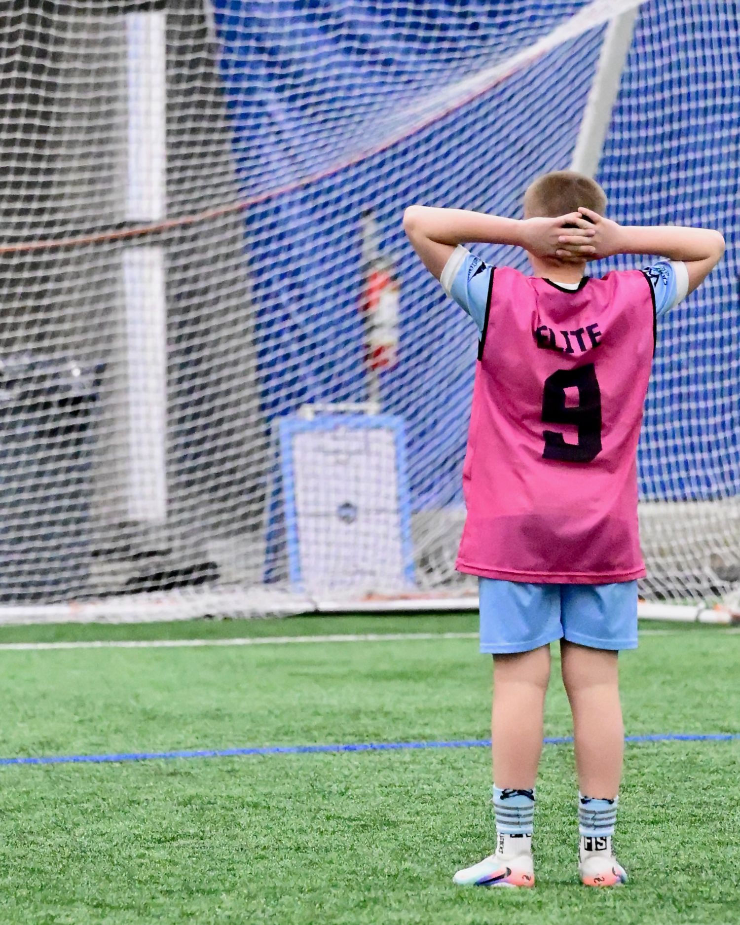 Youth soccer player number 9 in pink jersey stands with hands on head near goal.