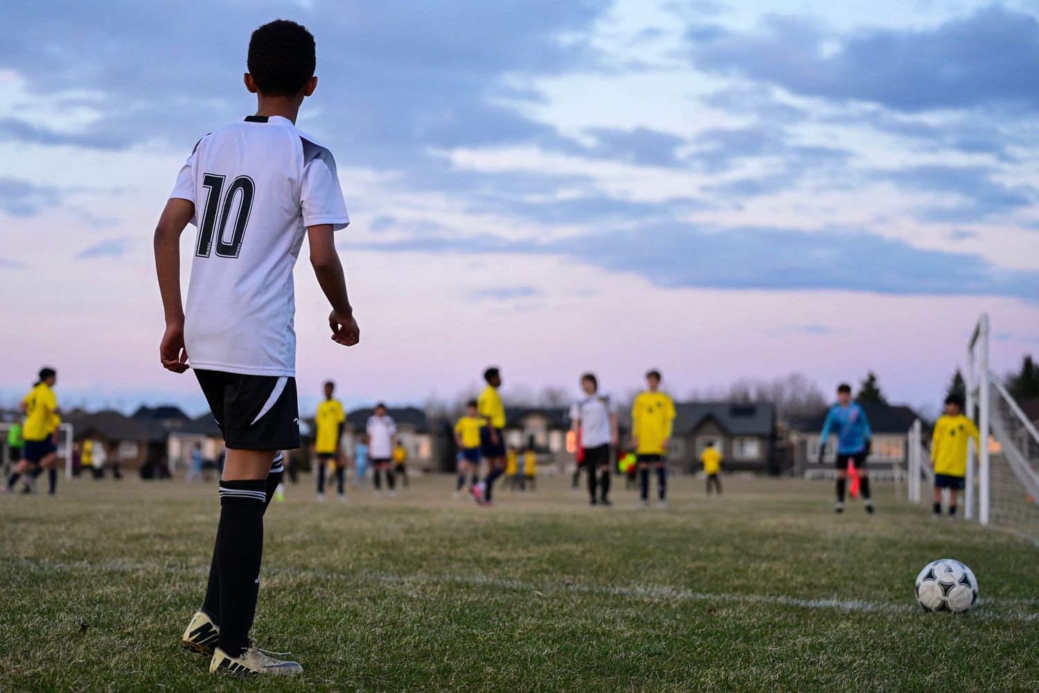 A youth soccer player wearing number 10 stands on a field at dusk watching teammates in the distance.