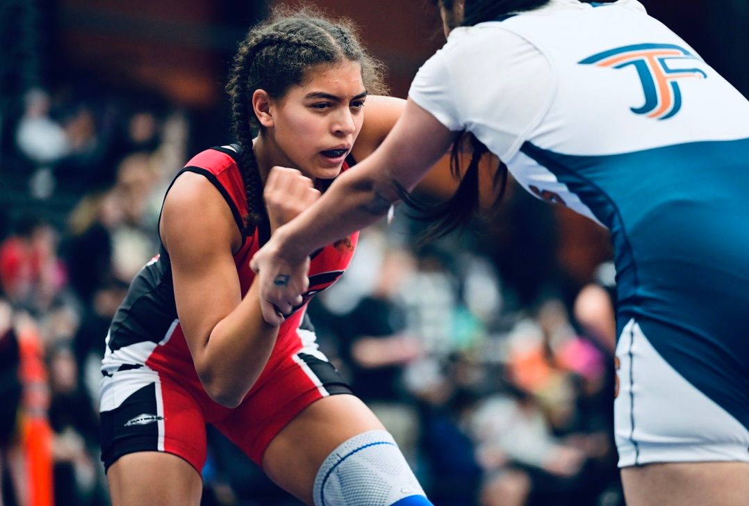 Two female wrestlers compete intensely in a grappling match wearing red and blue singlets.