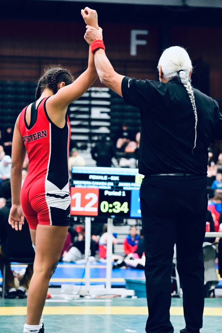 A female wrestler in a red singlet has her arm raised by a referee after winning a match.