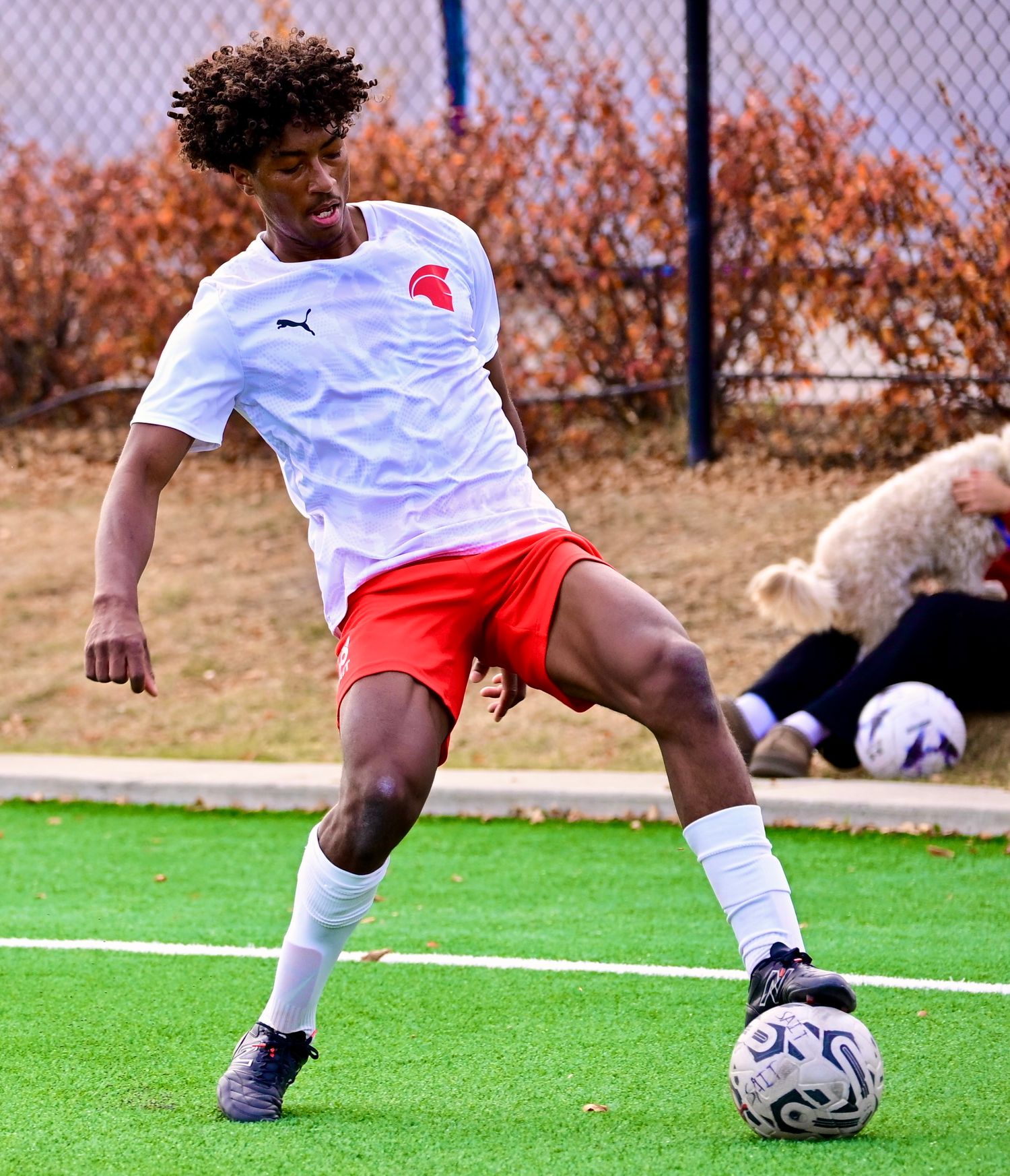 A young male soccer player in white and red Puma kit dribbles a ball on an outdoor turf field.