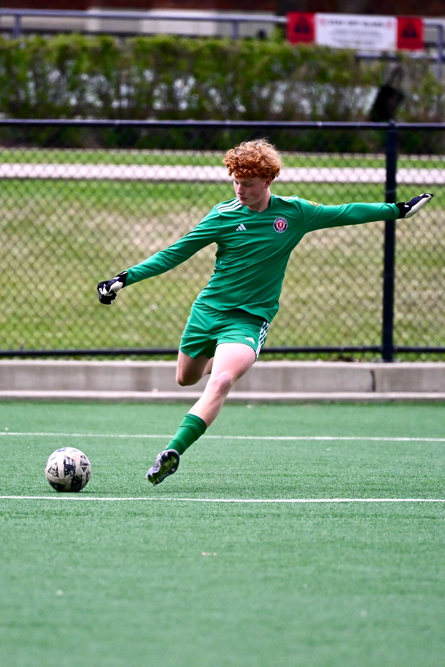 Young soccer goalkeeper in green Adidas kit preparing to kick ball on turf field.