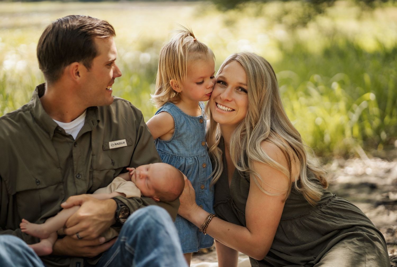A happy family of four sits outdoors, parents smiling with a toddler girl and newborn baby in a sunny green setting.