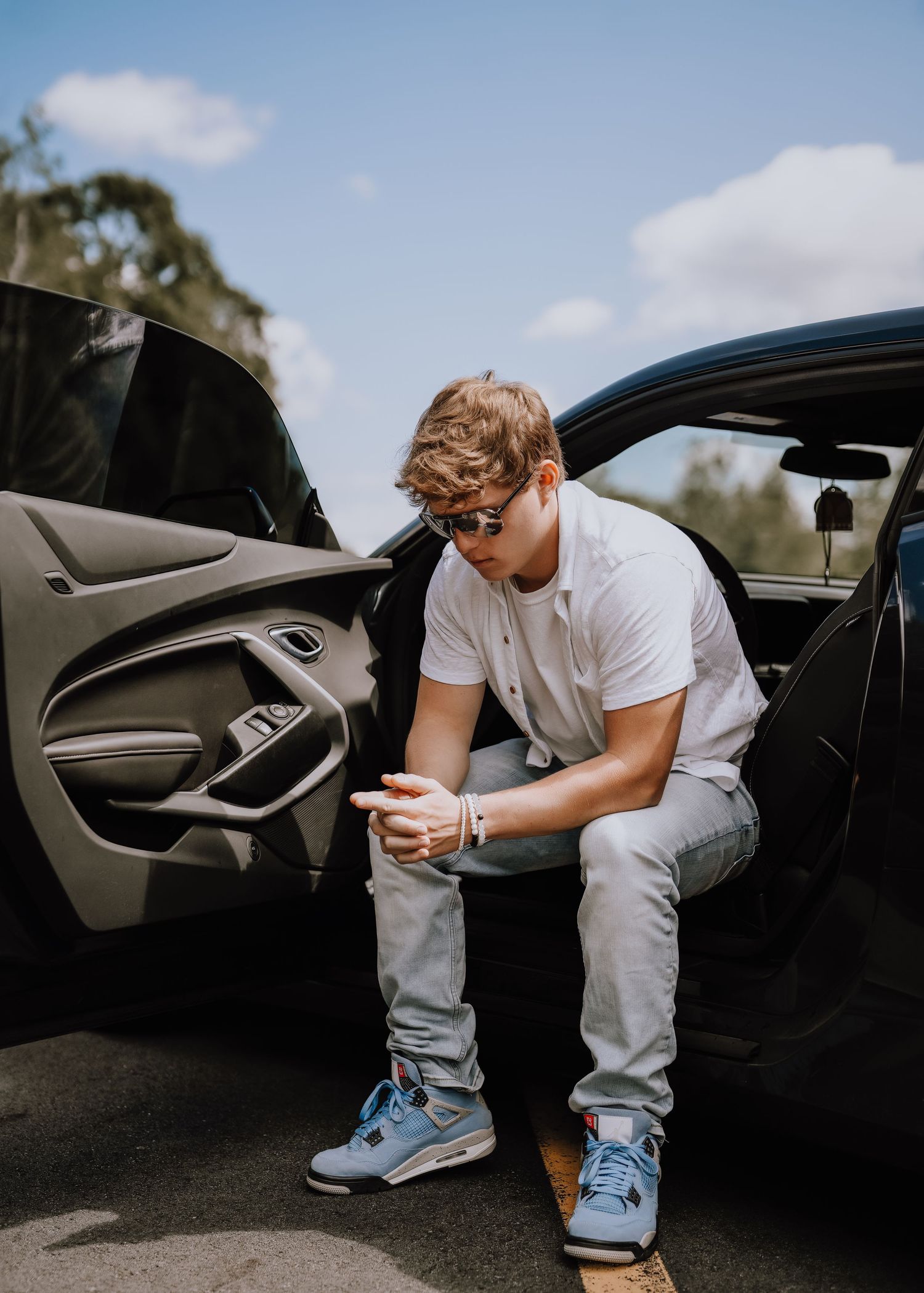 Young man in white polo, gray jeans, and blue Jordan 4s sitting in car door, looking down at phone.