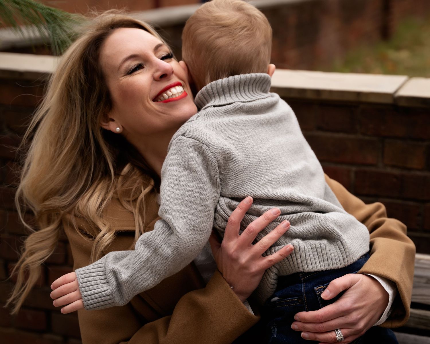 A smiling blonde woman in a camel coat lovingly embraces her young toddler son during a family portrait session.