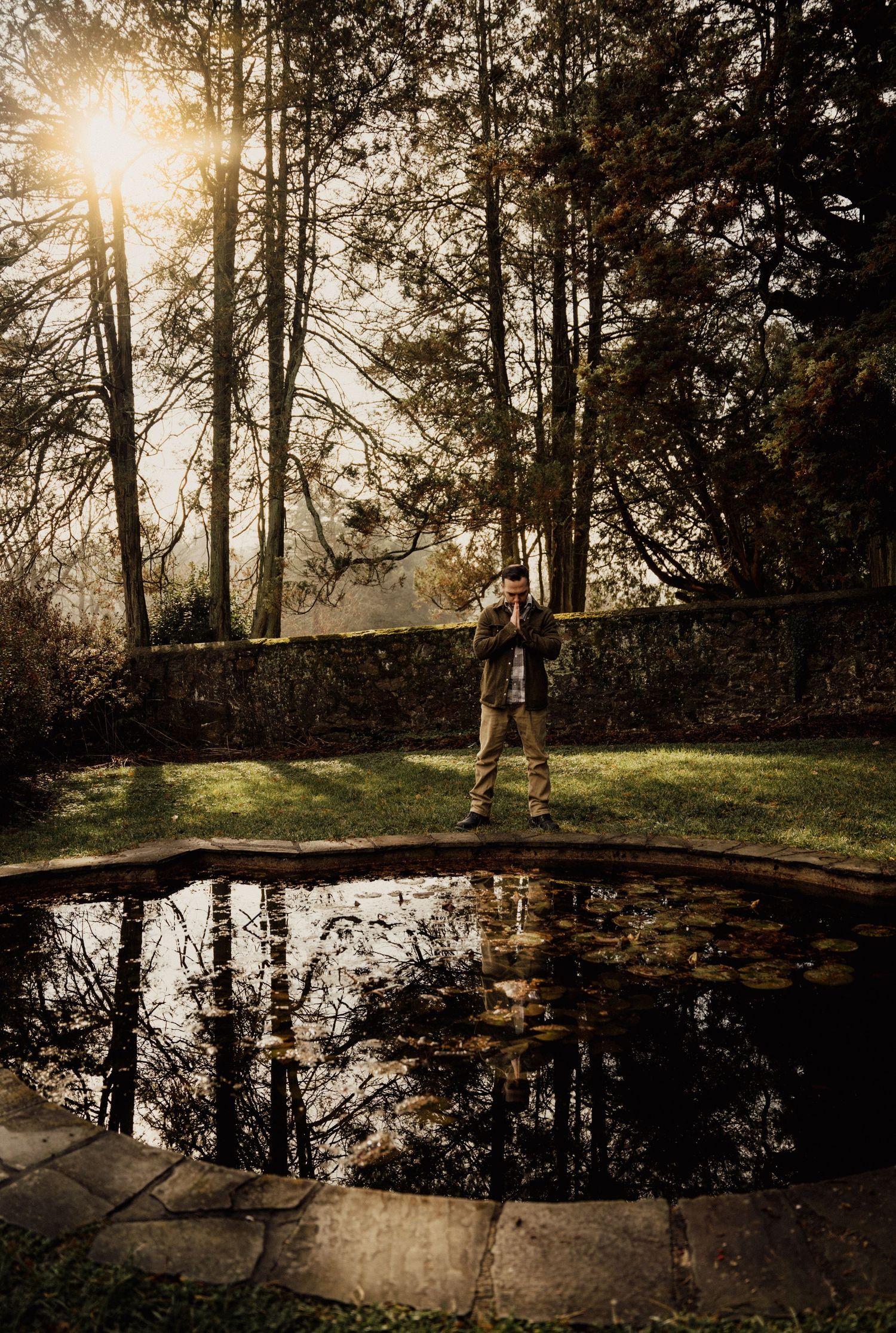 Man standing beside a reflective garden pond surrounded by autumn trees with golden sunlight filtering through the branches.