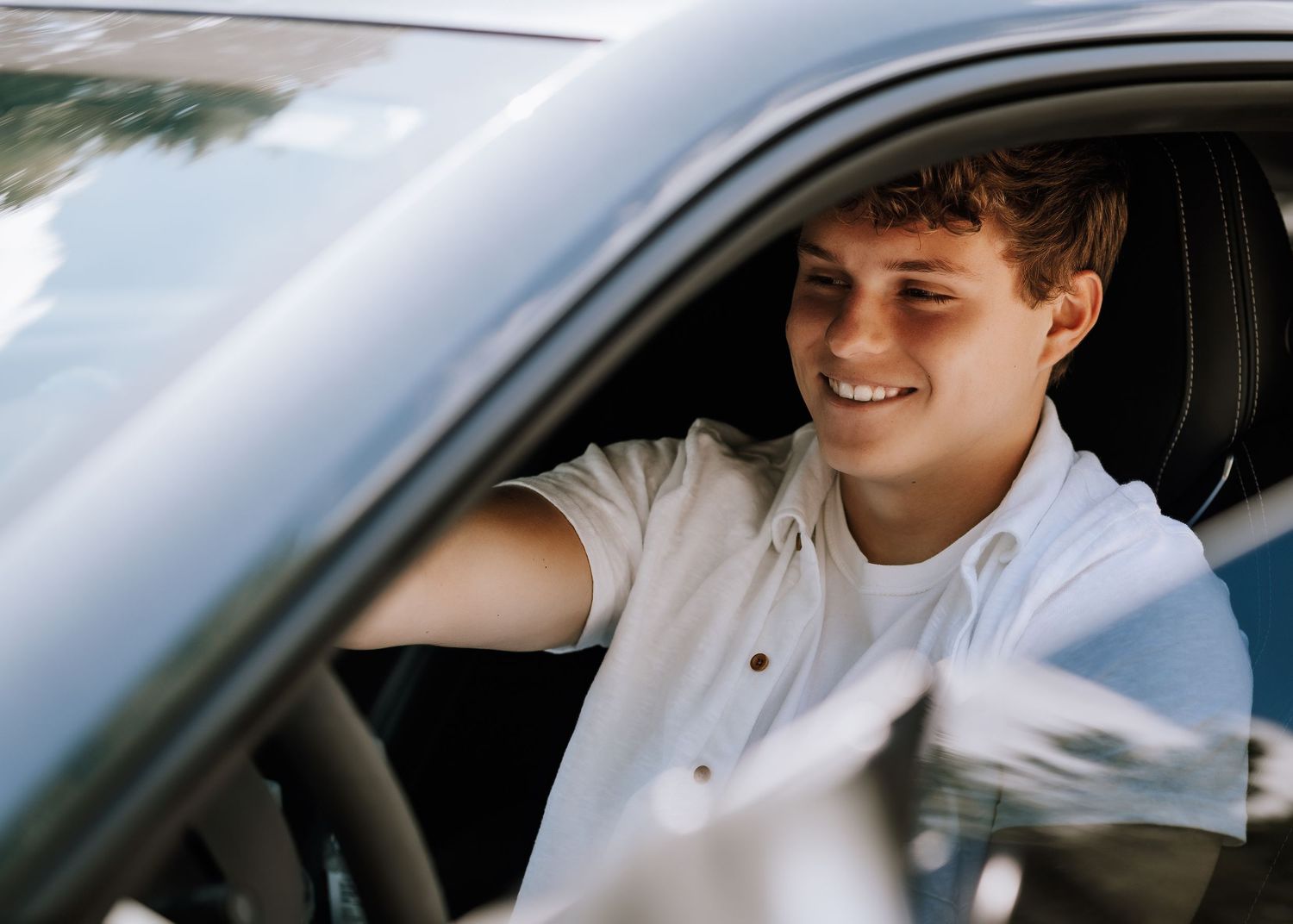 Smiling young man sitting in driver seat of car, wearing white shirt, enjoying a sunny day drive.