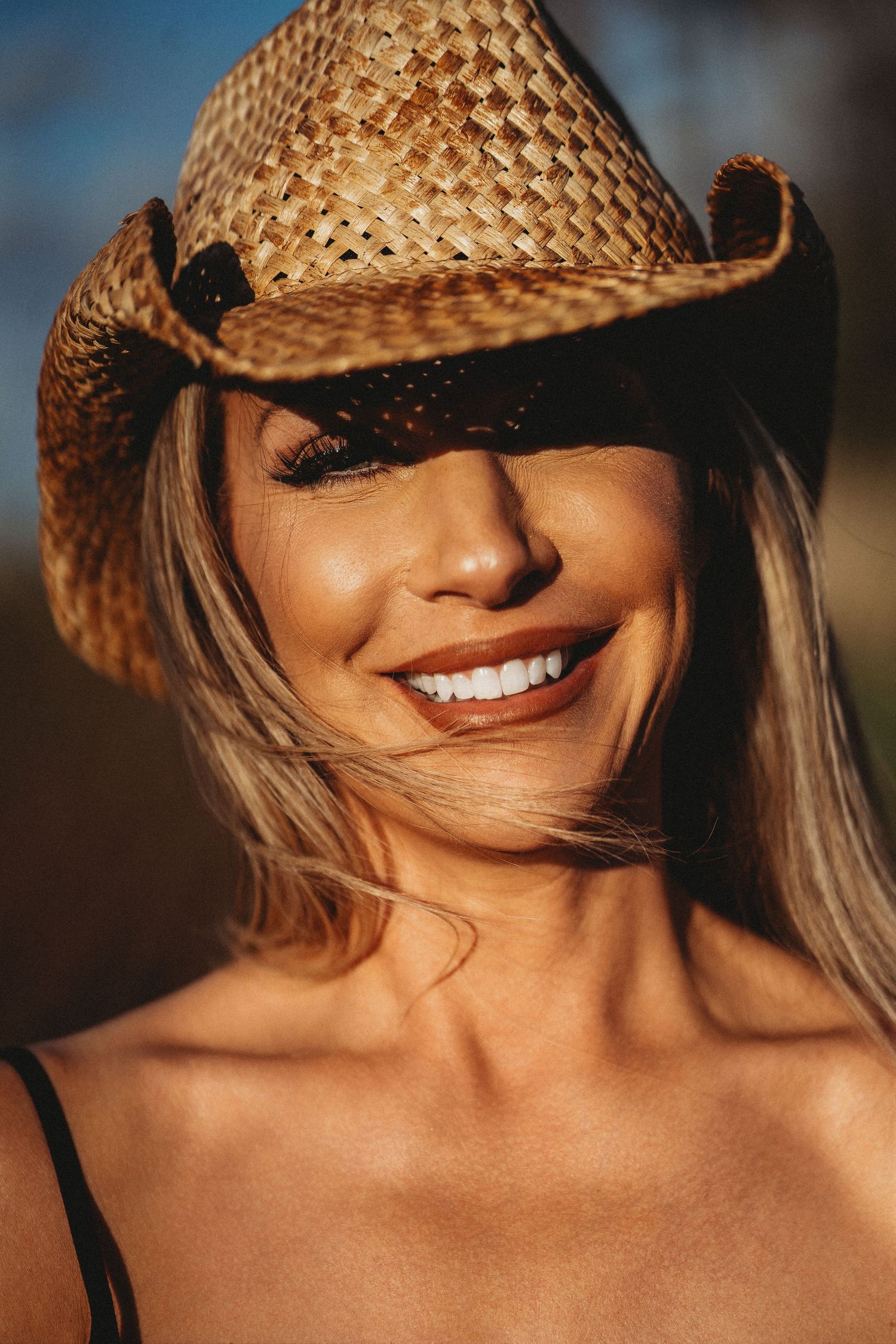 Smiling blonde woman wearing a woven straw cowboy hat tilted over her eyes, photographed outdoors in warm sunlight.