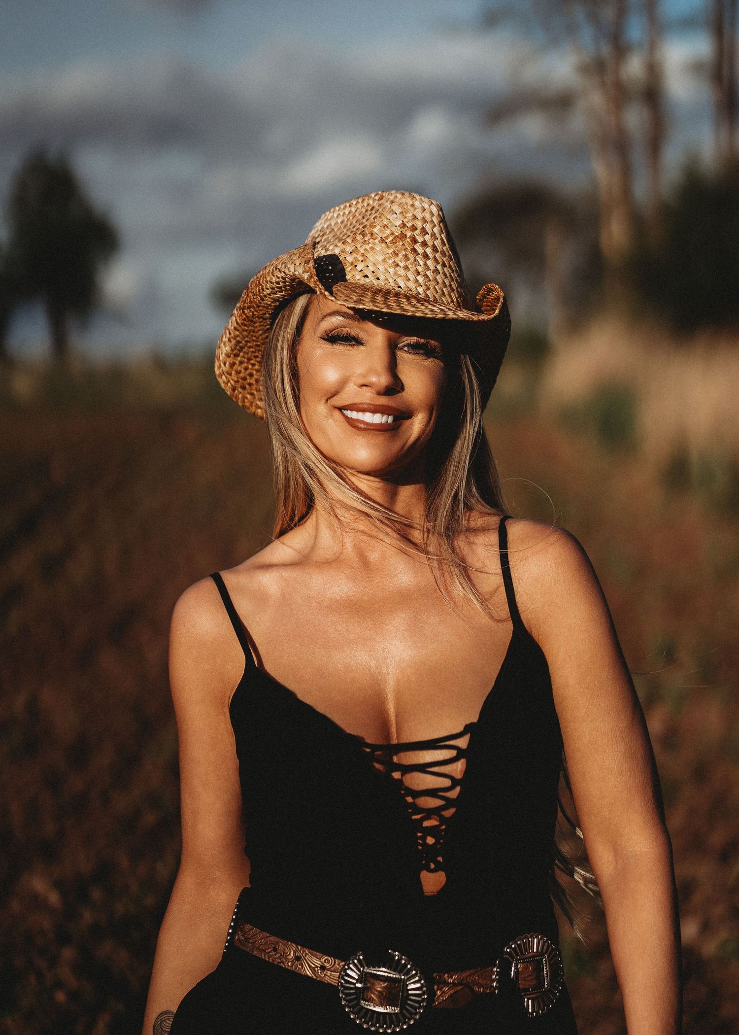 Smiling blonde woman wearing a straw cowboy hat and black lace-up bodysuit with a western belt outdoors.