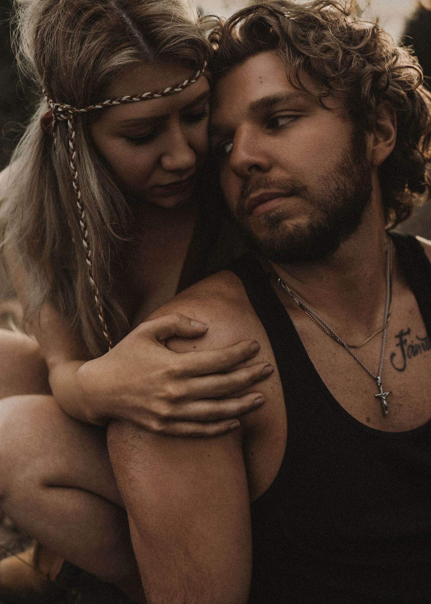 A bohemian couple embraces tenderly, woman in braided headband resting on bearded mans shoulder outdoors.