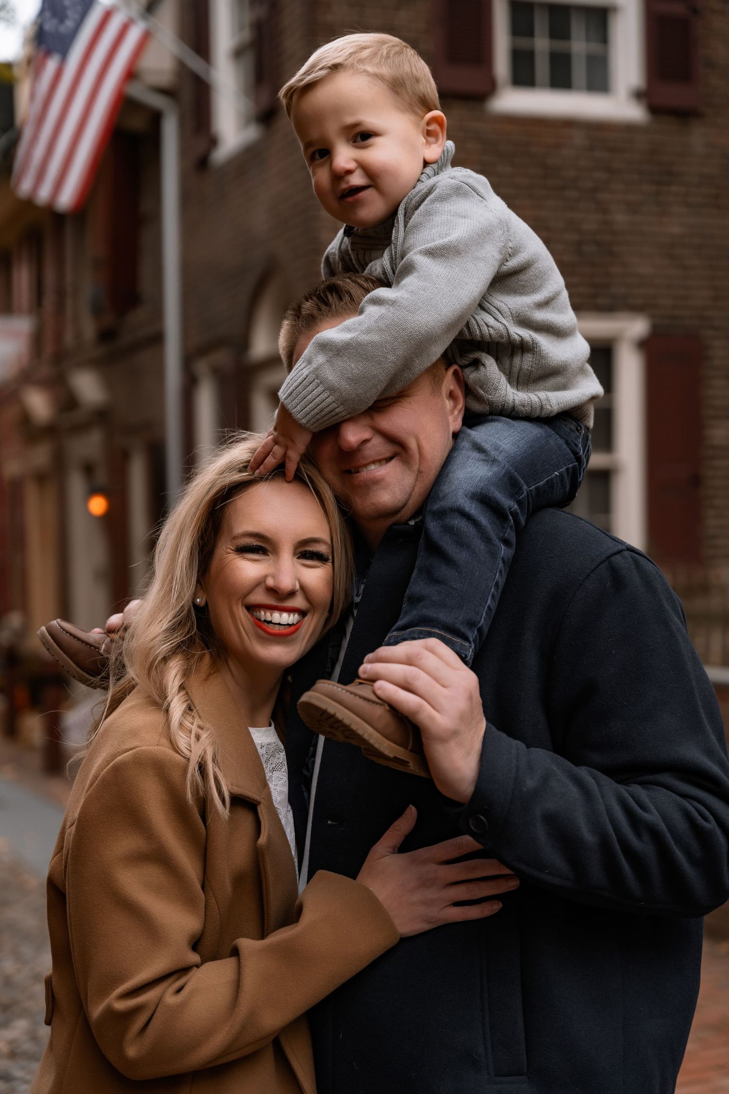 A happy family poses on a historic brick street, with a young boy sitting on his father's shoulders covering dad's eyes.