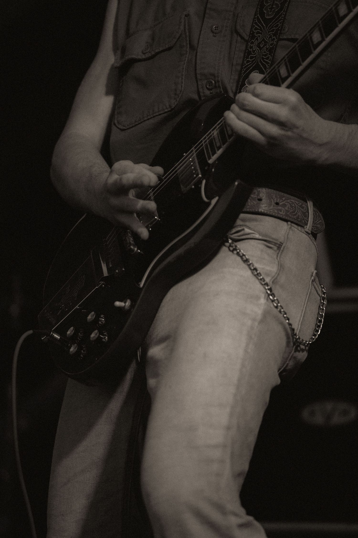 A black and white close-up of a guitarist playing a dark electric guitar, wearing jeans with a chain belt.