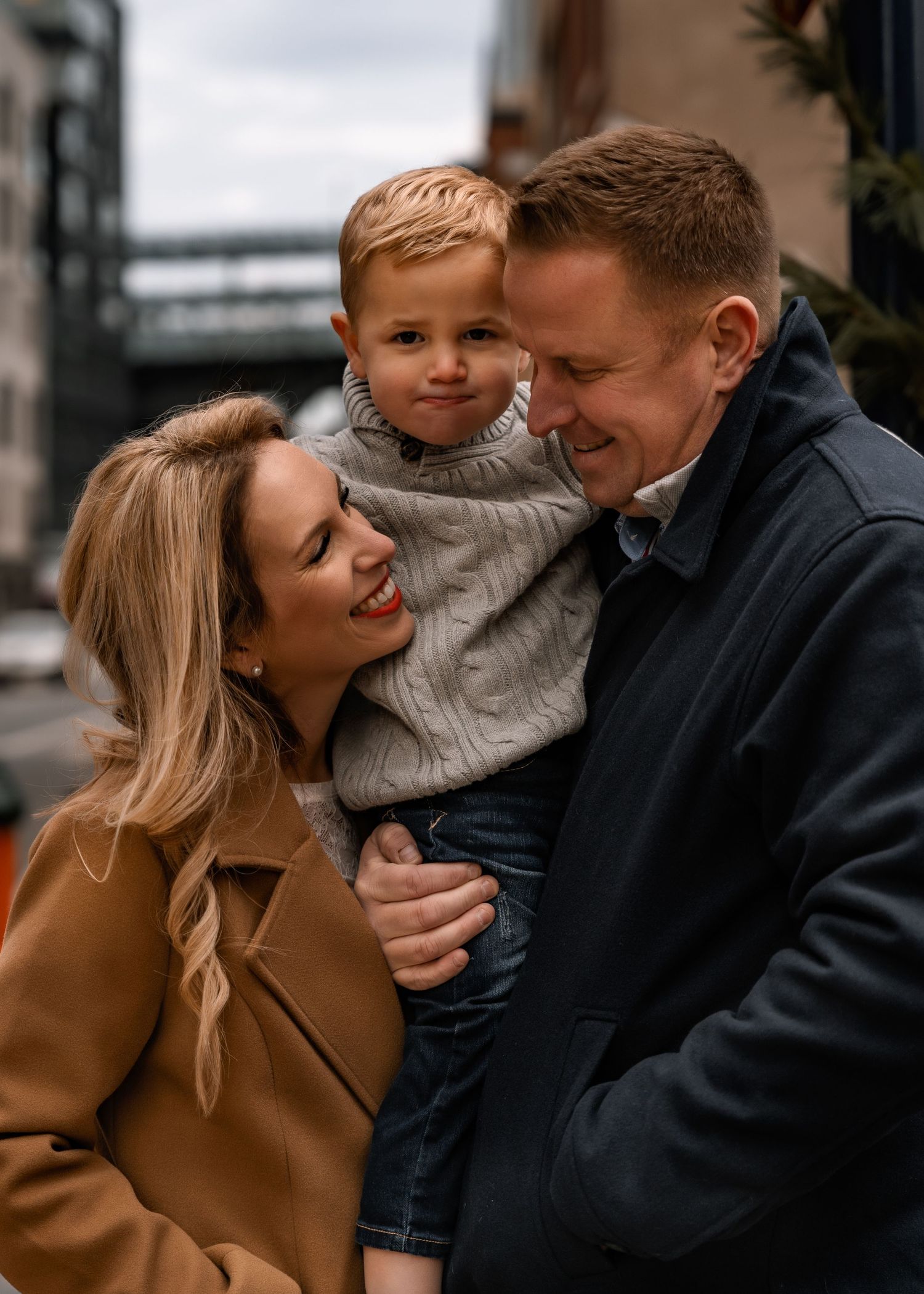 Happy family portrait with parents and young son embracing on urban street in winter coats.