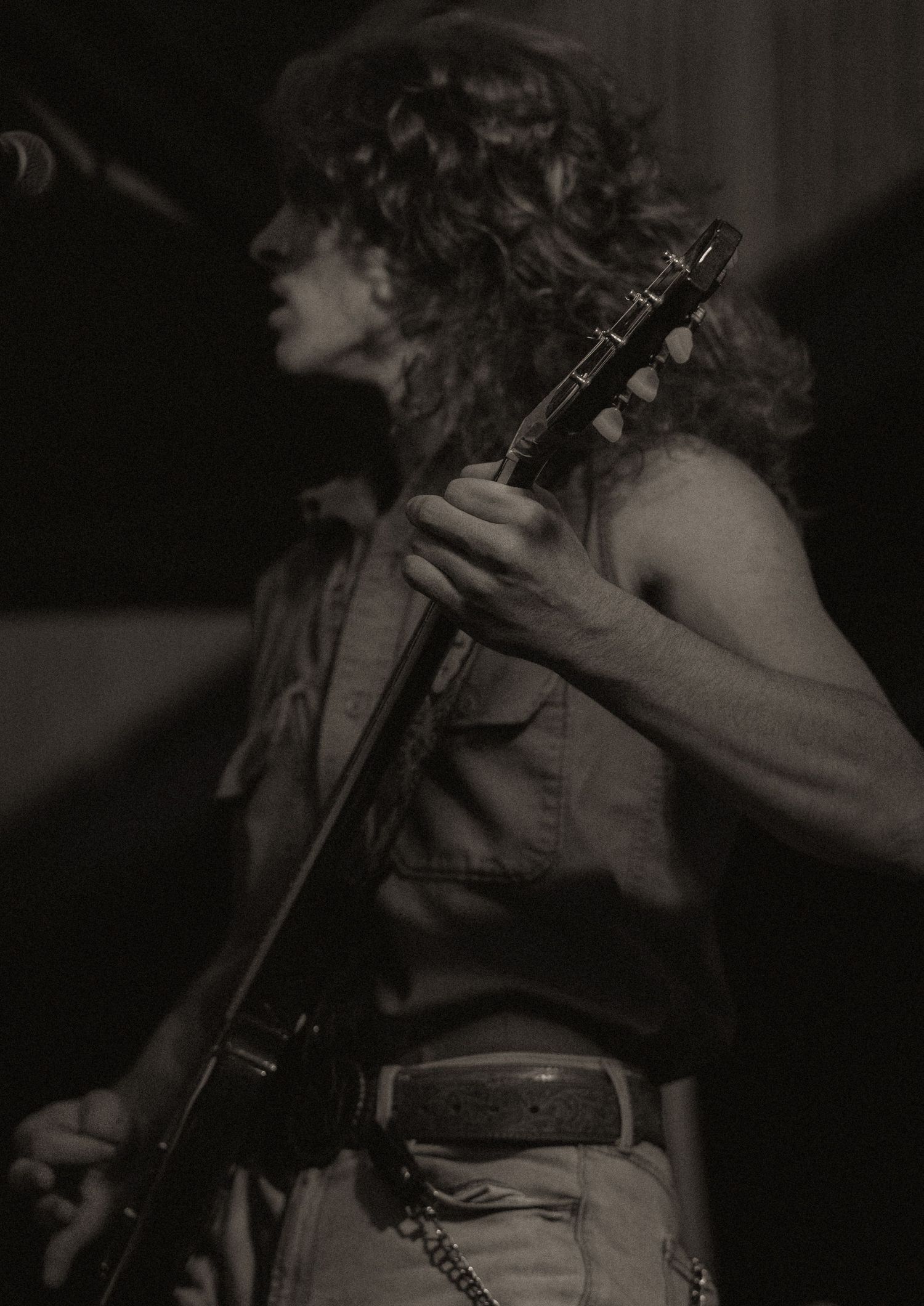 A black and white side profile of a curly-haired guitarist playing an electric guitar on stage.