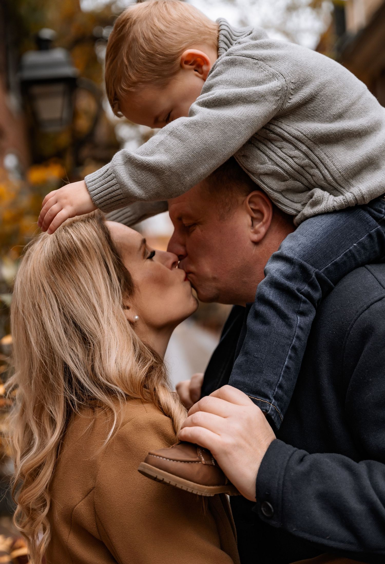 A couple shares a kiss while their toddler boy perches on dad's shoulders during an autumn family photo session.