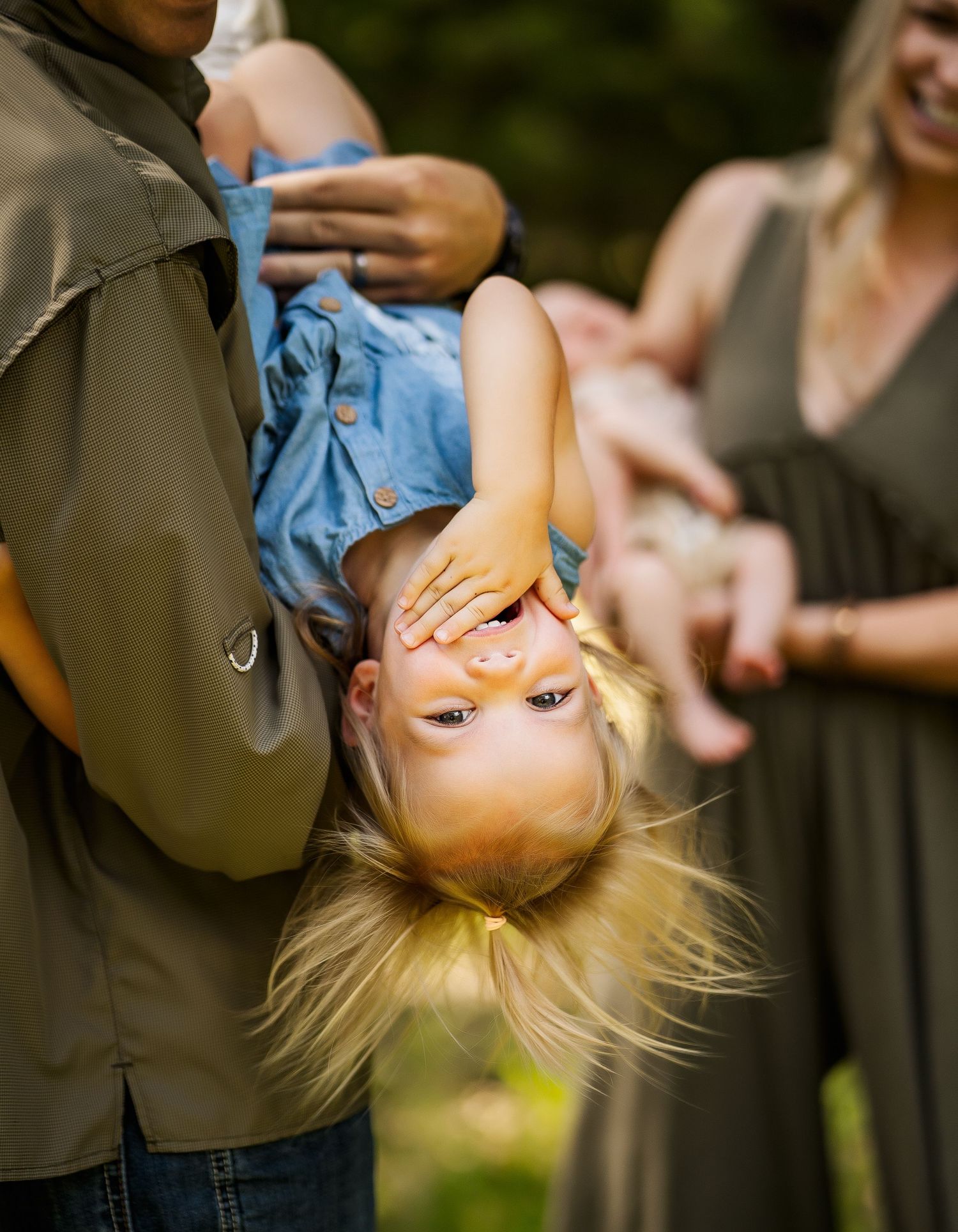A laughing toddler held upside down by a parent outdoors, with a woman holding a baby in the background.