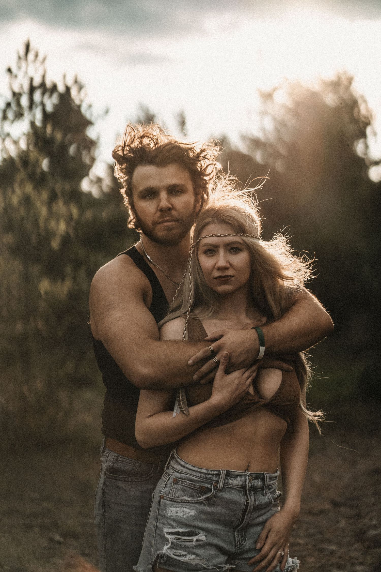 Bohemian couple embracing outdoors at sunset, man in tank top hugging woman in boho headband and denim shorts.