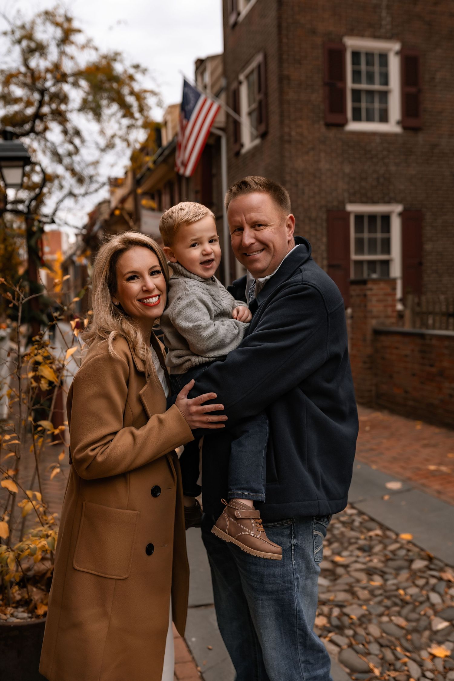 A cheerful family of three poses on a tree-lined sidewalk with fall foliage and an American flag in the background.