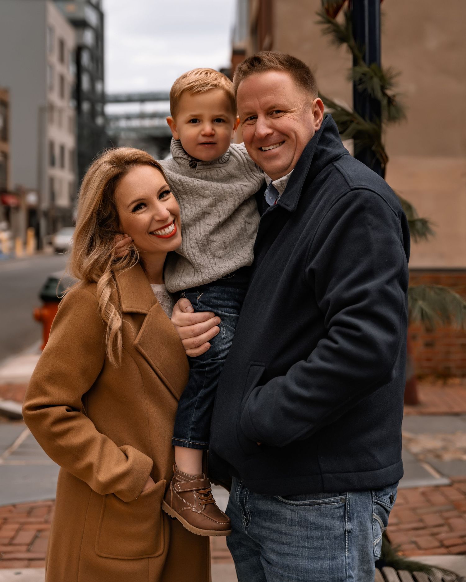 A happy family of three poses on a city street, with dad holding their smiling toddler son in a cozy fall setting.