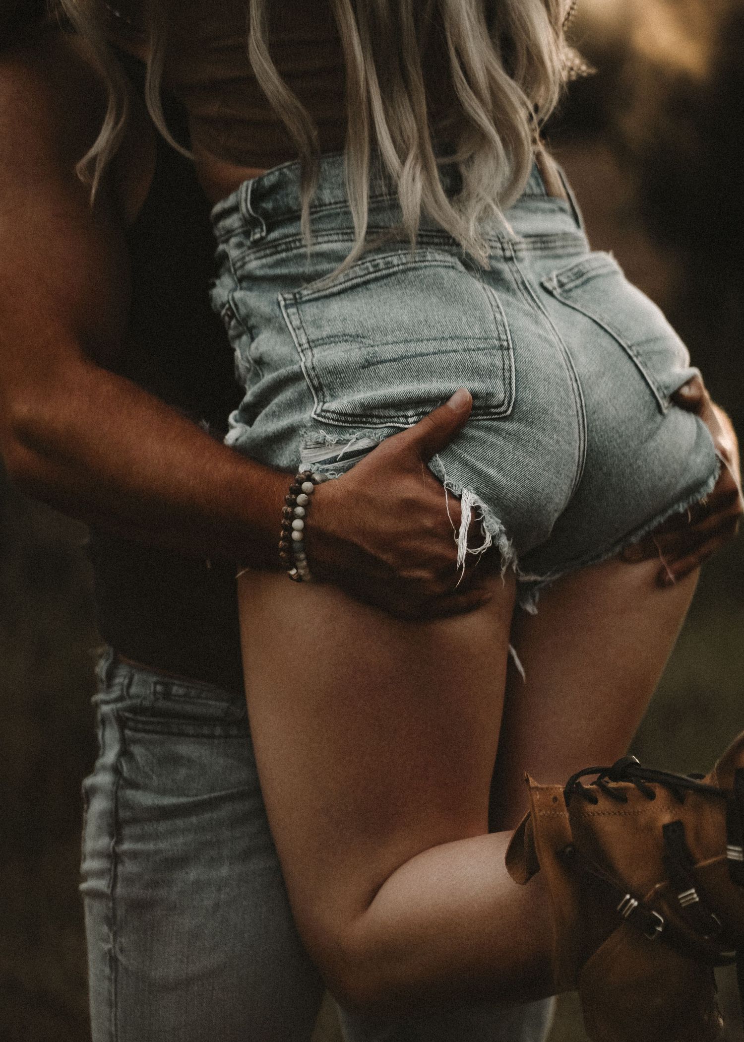 A couple embracing outdoors, man's hand on woman's hip wearing denim cutoff shorts, with a bracelet visible.