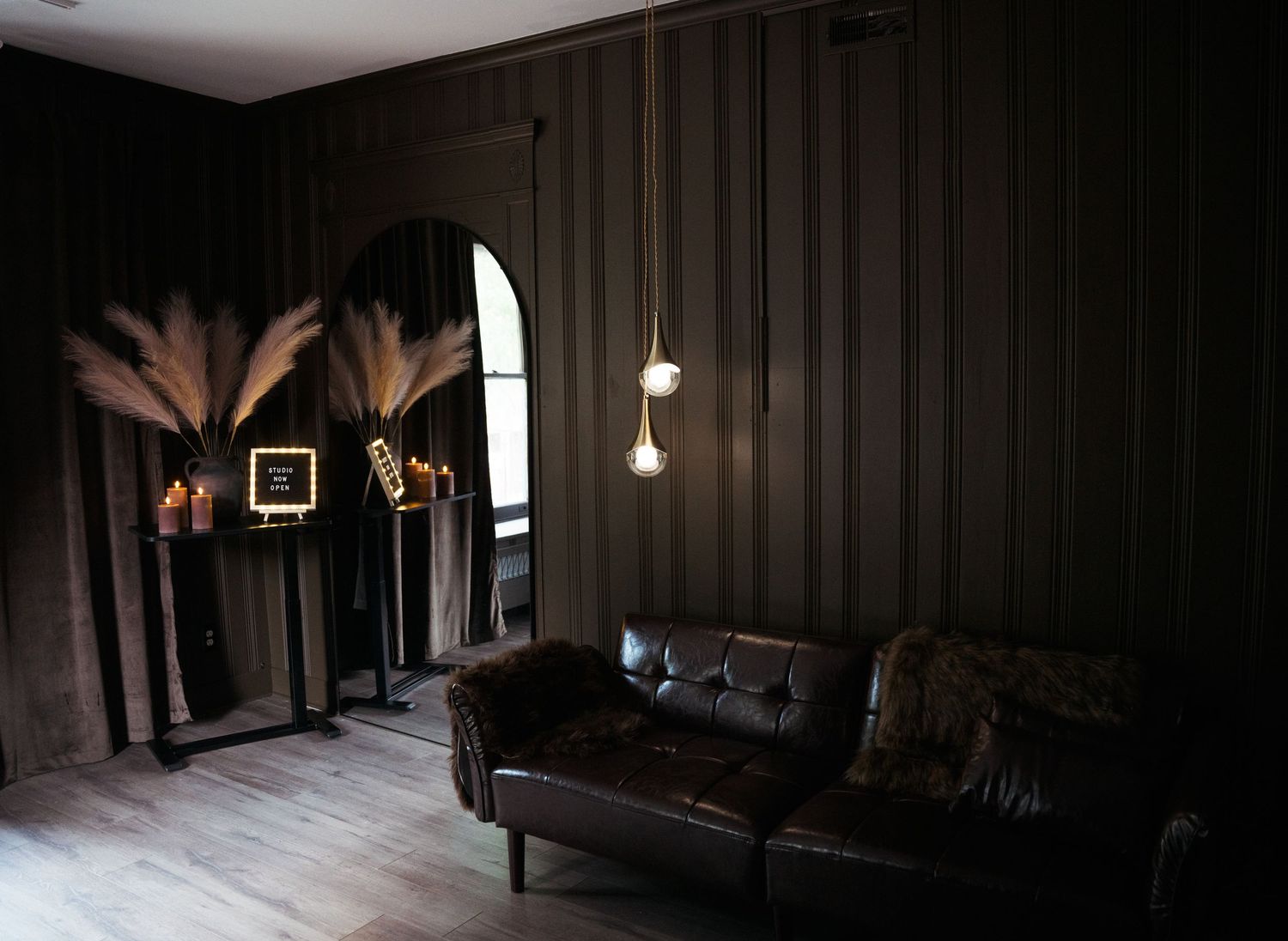 Dark moody lounge with tufted leather sofa, pendant lights, arched mirror, and pampas grass decor against dark paneled walls.