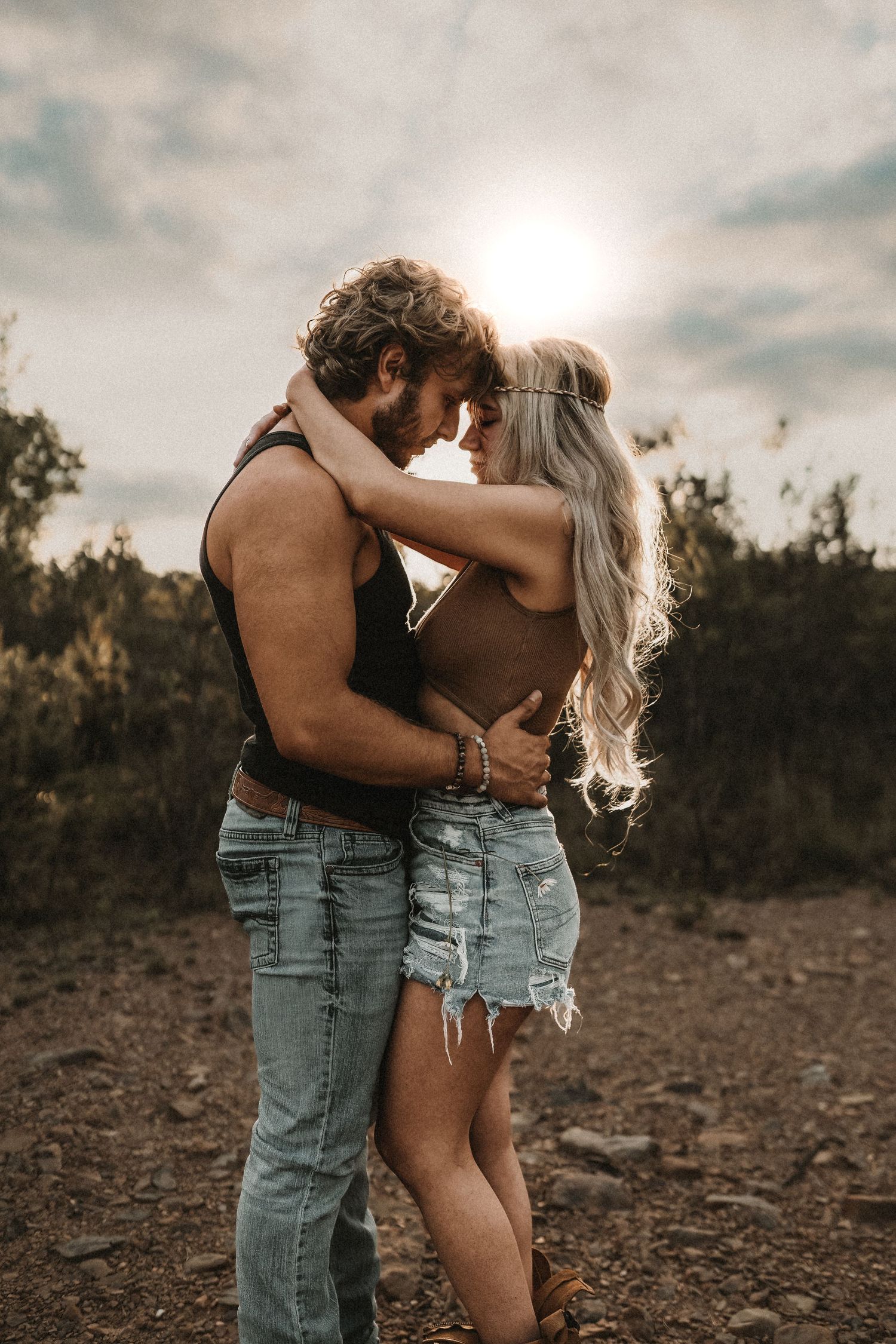 A couple embraces romantically at sunset in a rustic outdoor setting, with the woman in denim shorts and the man in jeans.