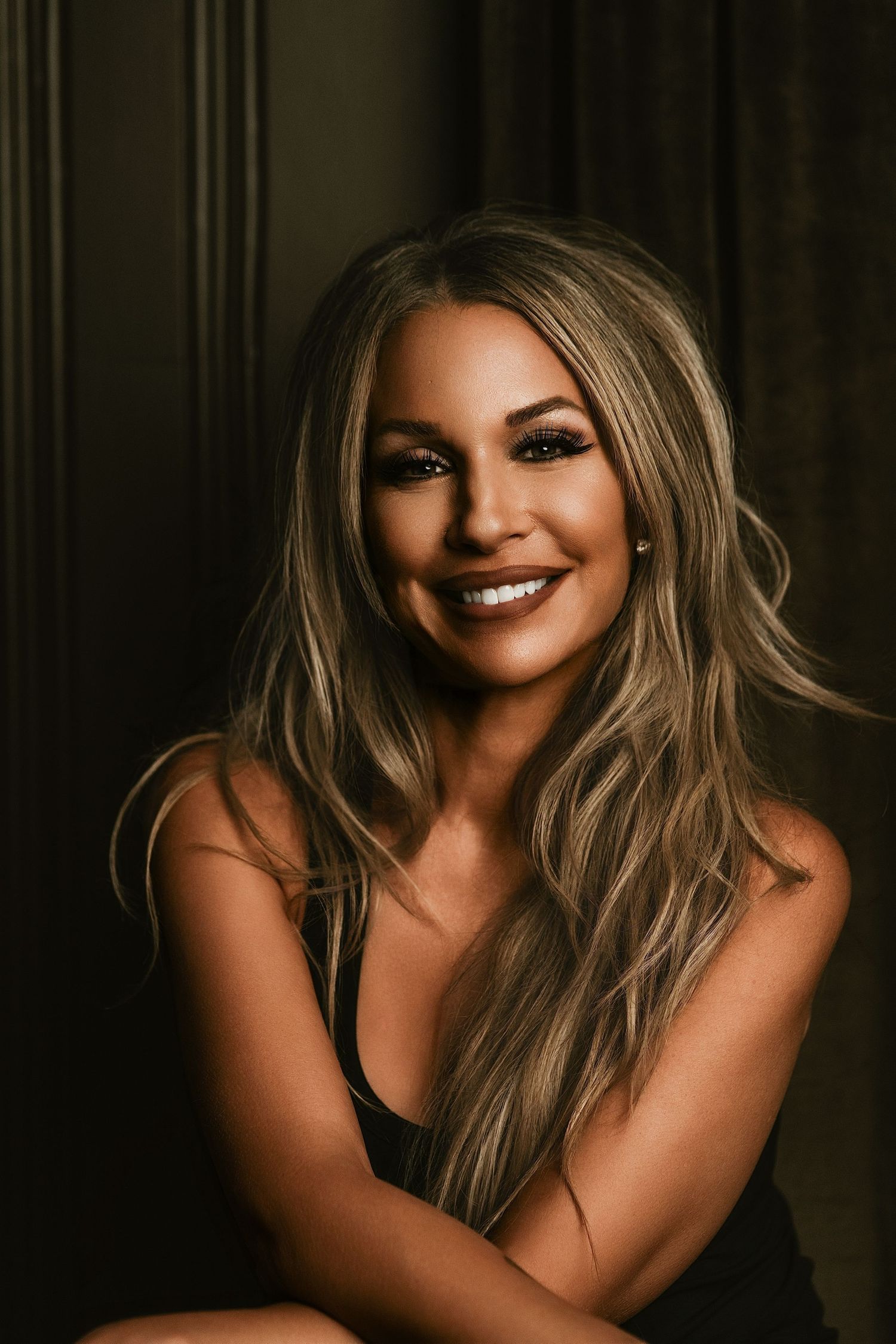Smiling blonde woman with long wavy hair poses confidently in a dark studio setting wearing a black top.