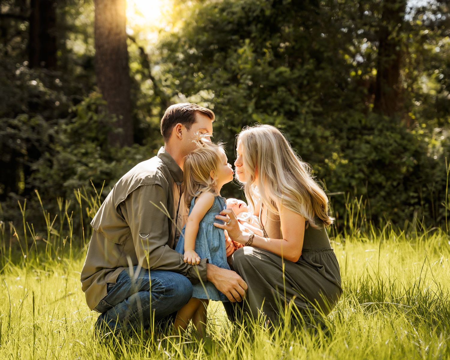 Family of four sharing tender moment together in sunlit green meadow surrounded by trees.