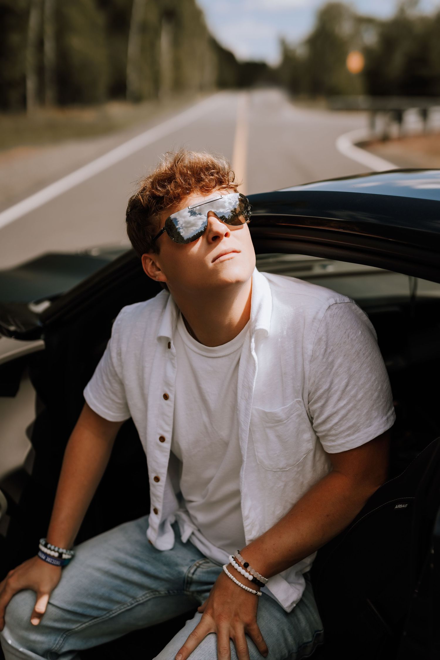 Young man in white shirt and reflective sunglasses leaning out of a black car on a scenic road.