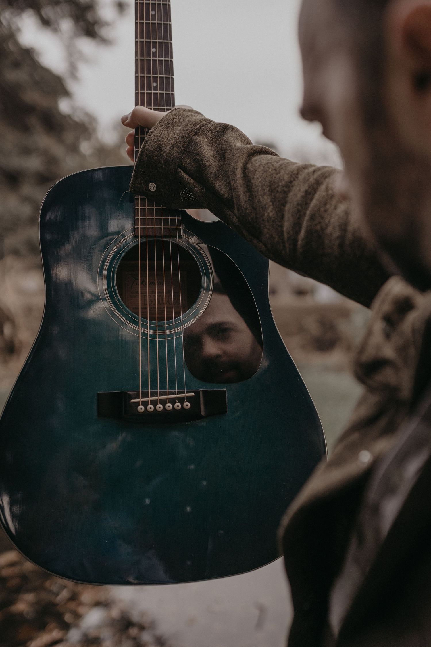 A bearded man holds a blue acoustic guitar up close to the camera, with his reflection visible in the body.