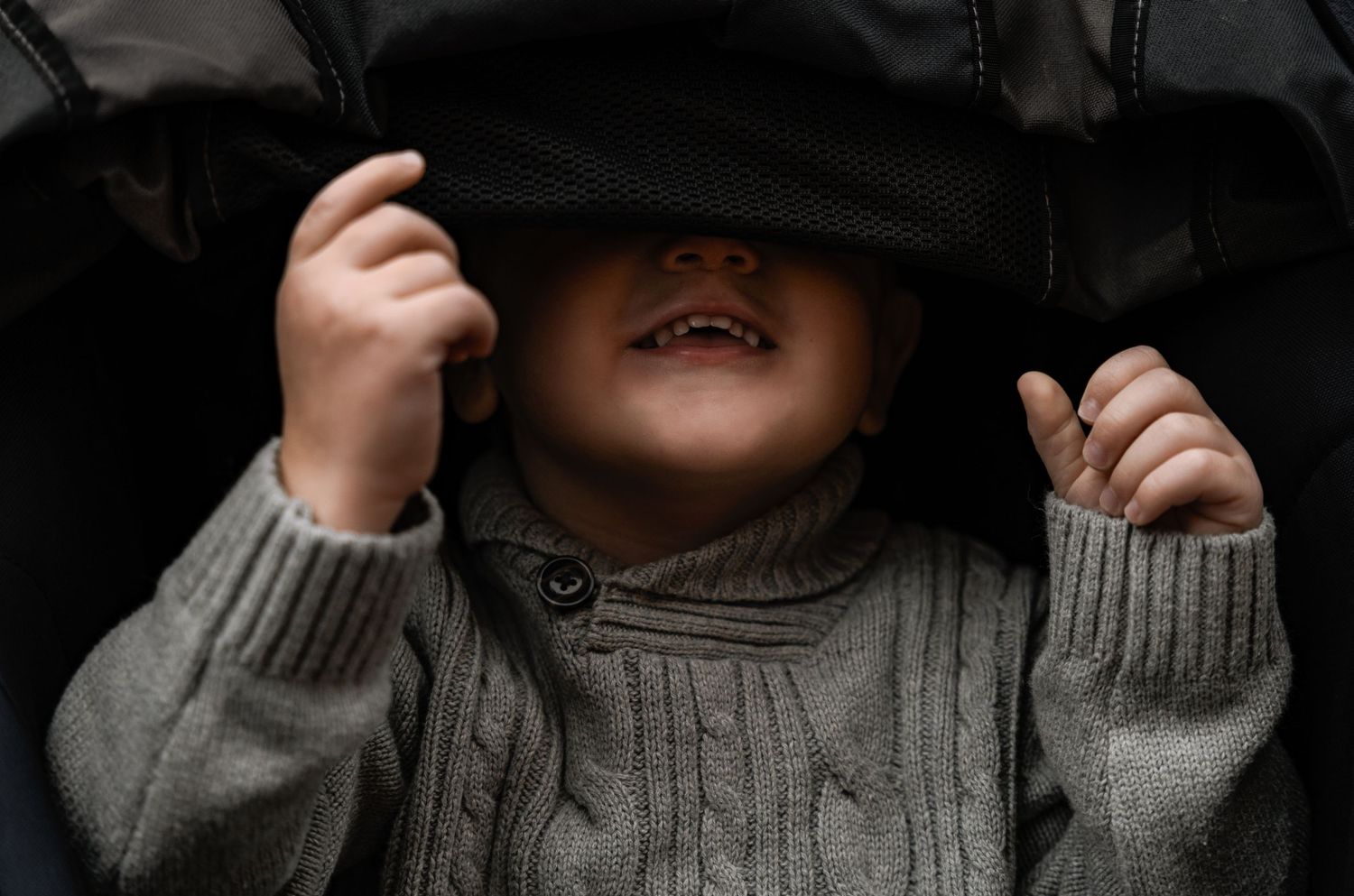 A playful toddler peeks out from under a dark hat, smiling with his gray cable-knit sweater visible below.