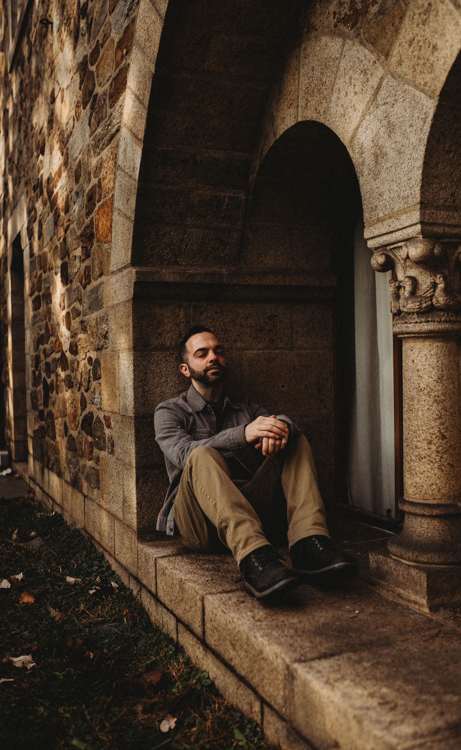 Bearded man sitting in stone archway of historic building with hands clasped together.