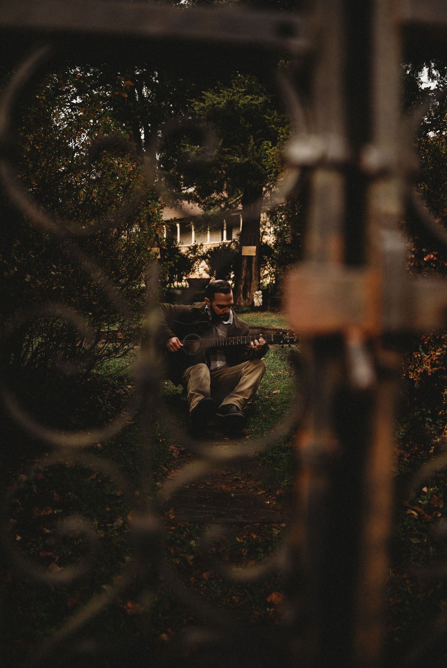 Man playing guitar outdoors, viewed through ornate iron gate with lush trees and white house in background.