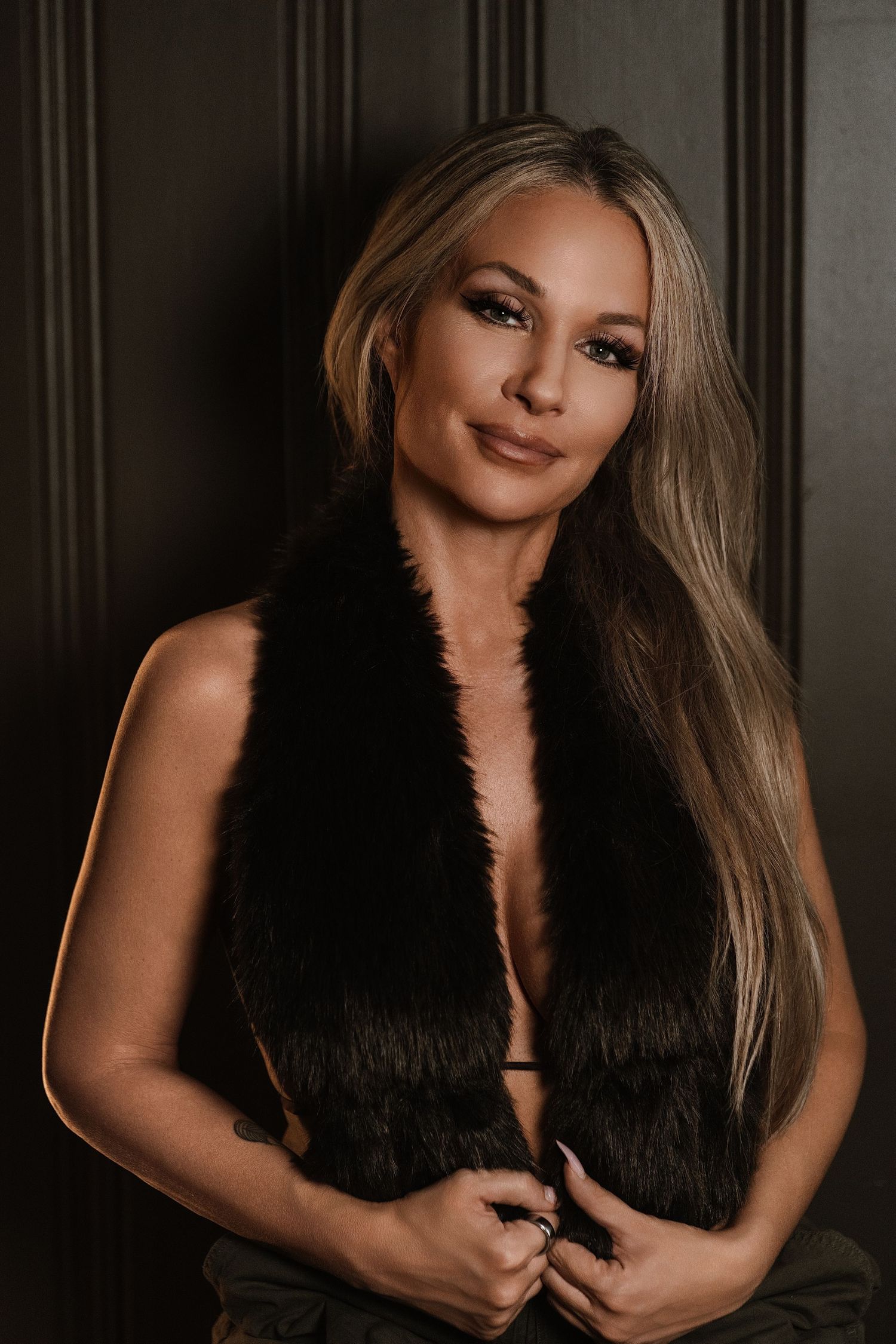 Blonde woman in black faux fur vest posing confidently against dark wood paneled wall with glamorous makeup.