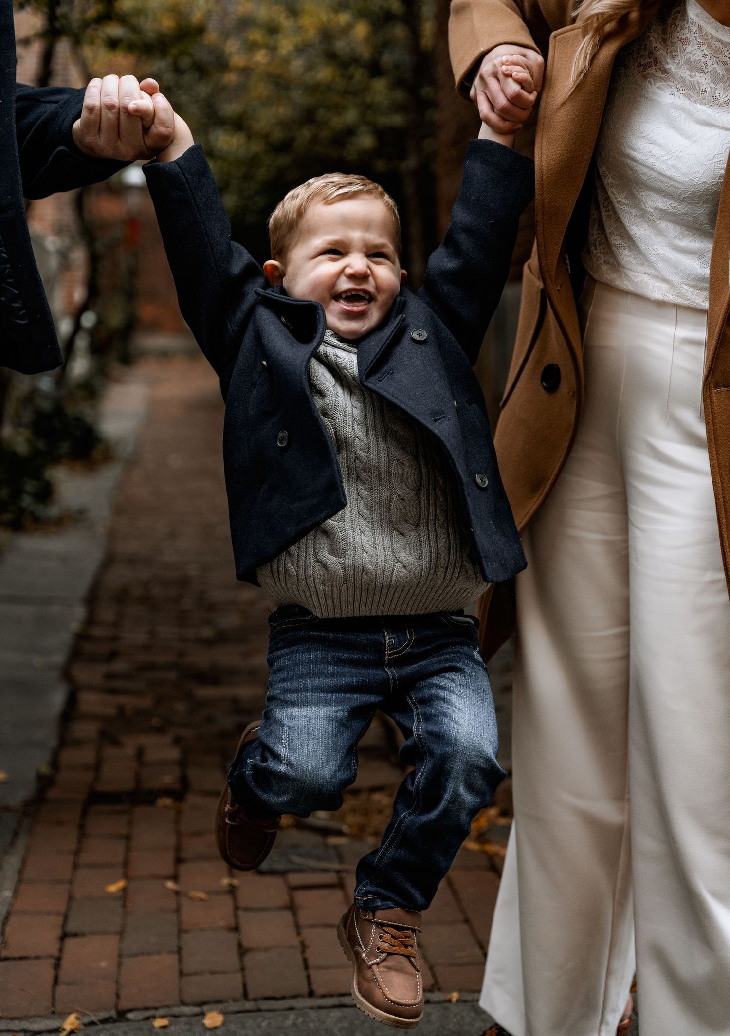 A joyful toddler boy jumps between his parents holding hands on a brick pathway during a fall family photoshoot.
