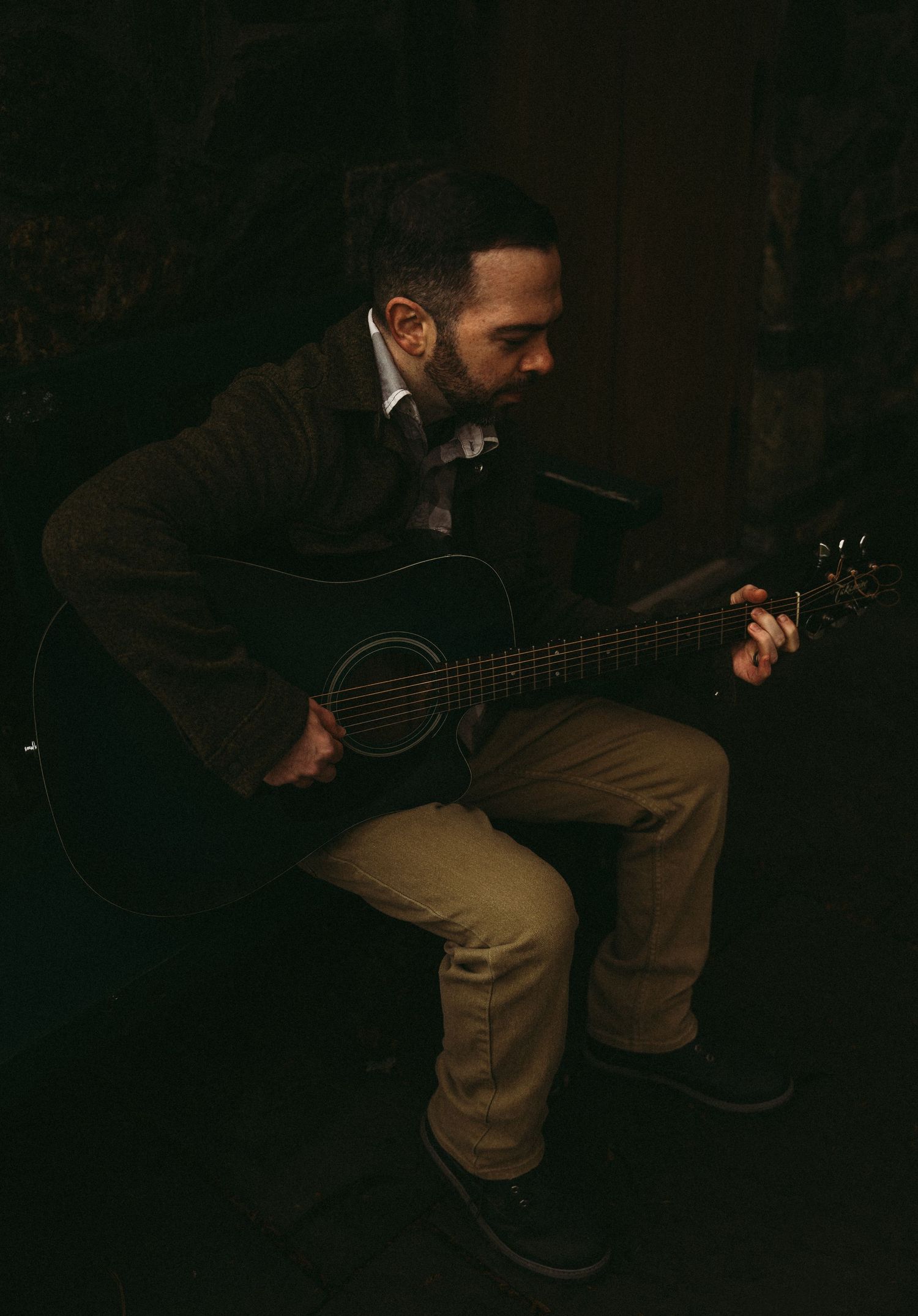Bearded musician playing acoustic guitar while seated in a dimly lit room wearing a dark blazer.