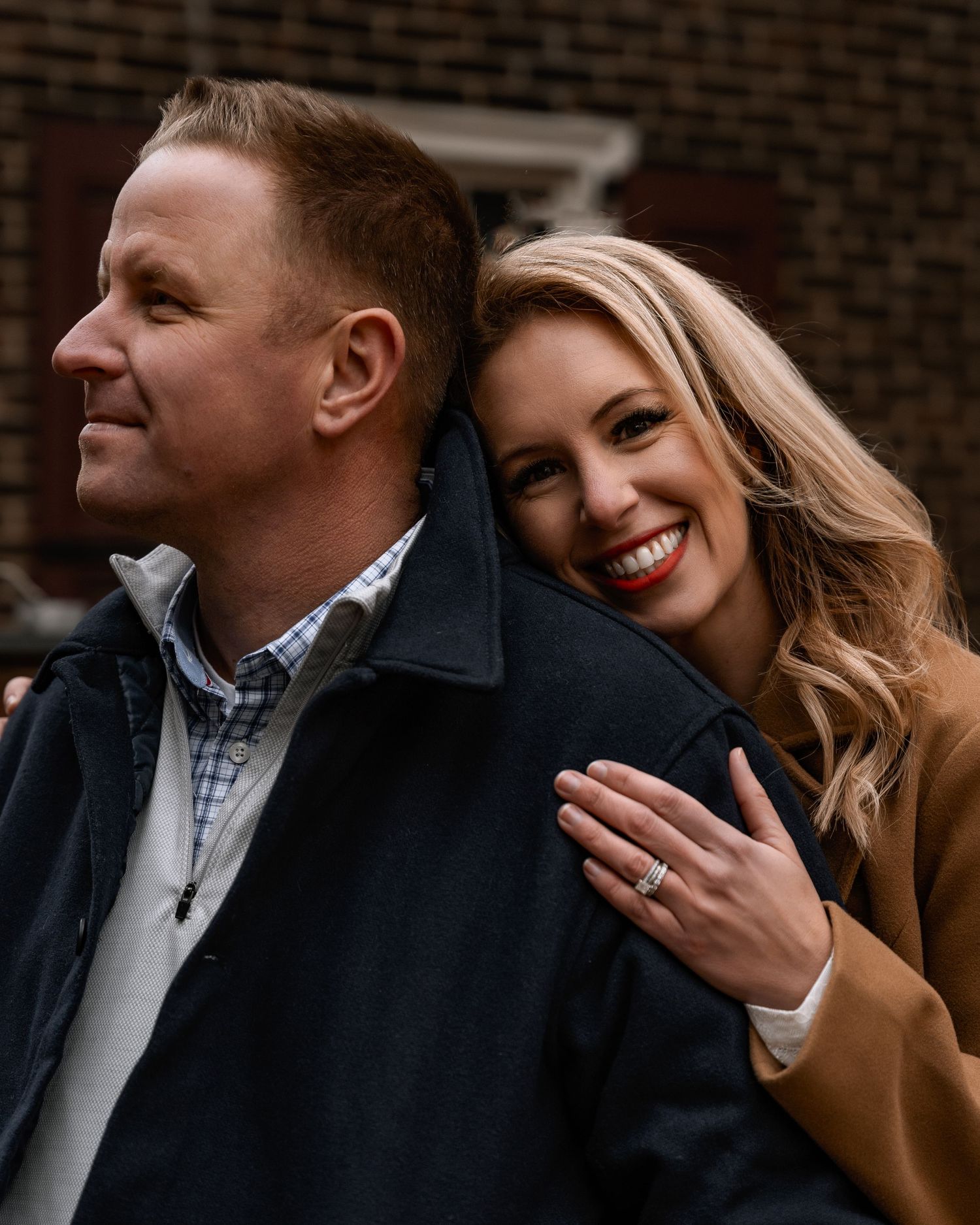 A smiling blonde woman leans against a man in a navy jacket, showing her engagement ring during a couples photo session.