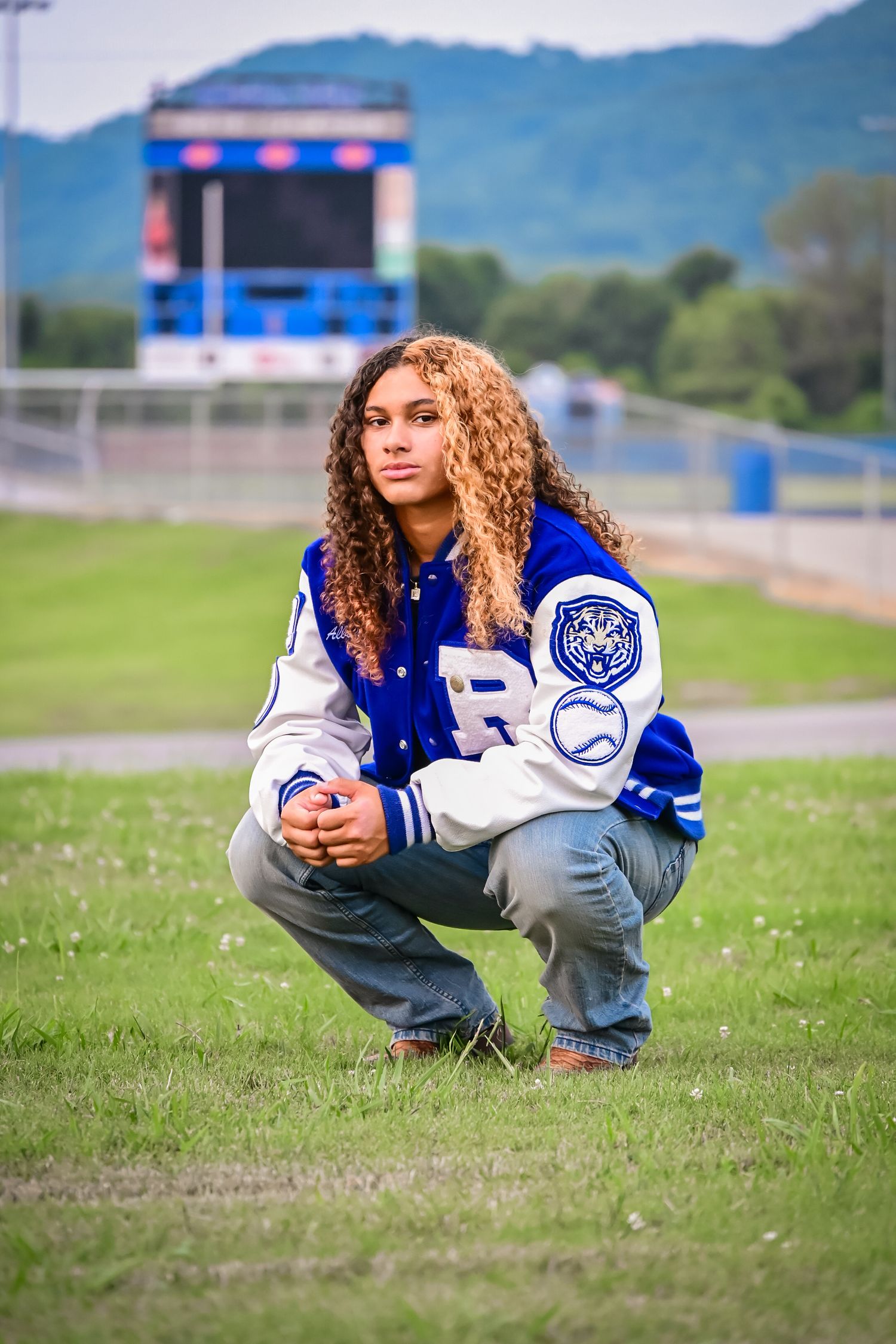 A young woman in a blue varsity jacket crouches on a grass field near a stadium scoreboard.
