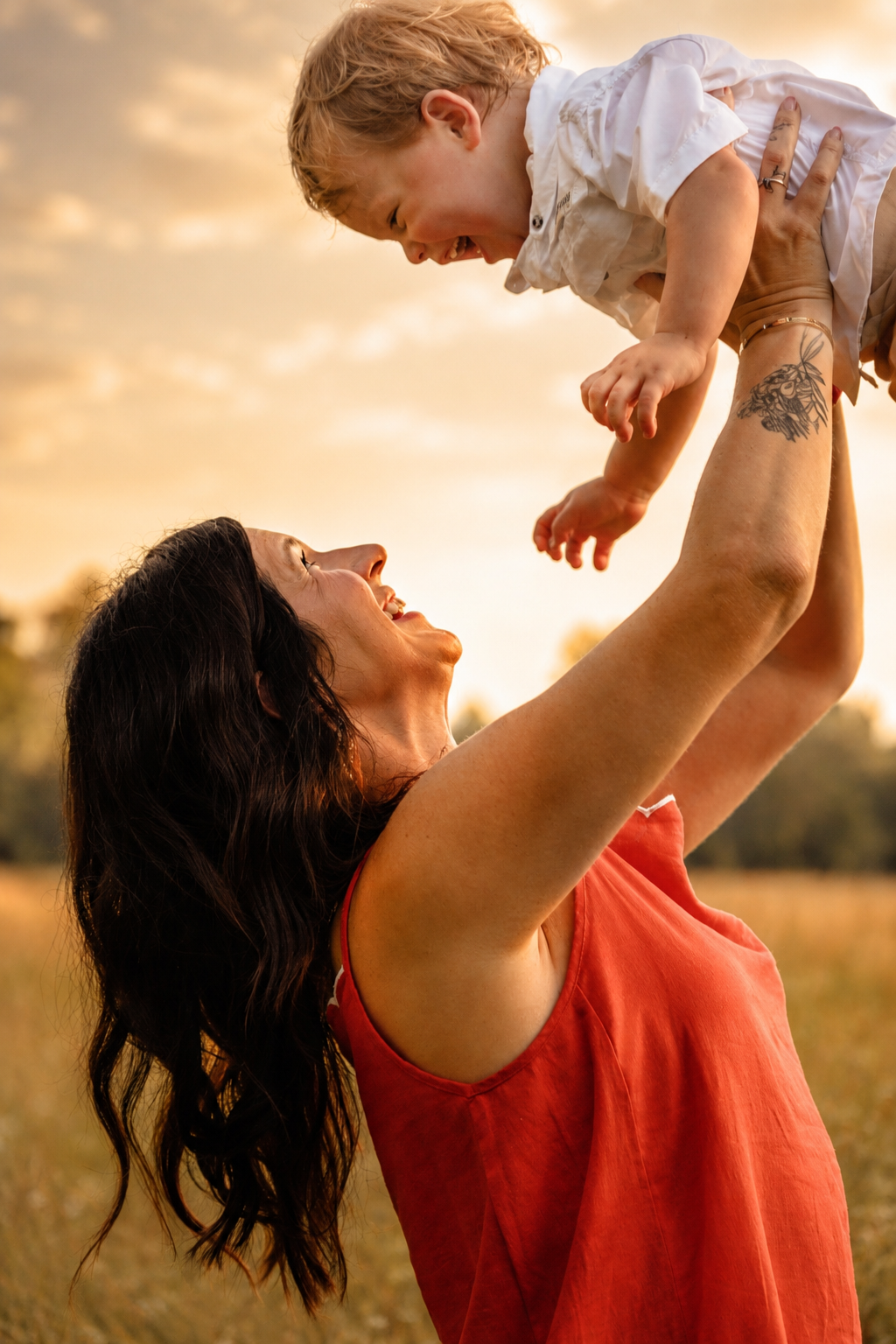 A joyful mother in a red dress lifts her laughing baby overhead in a golden sunset field.