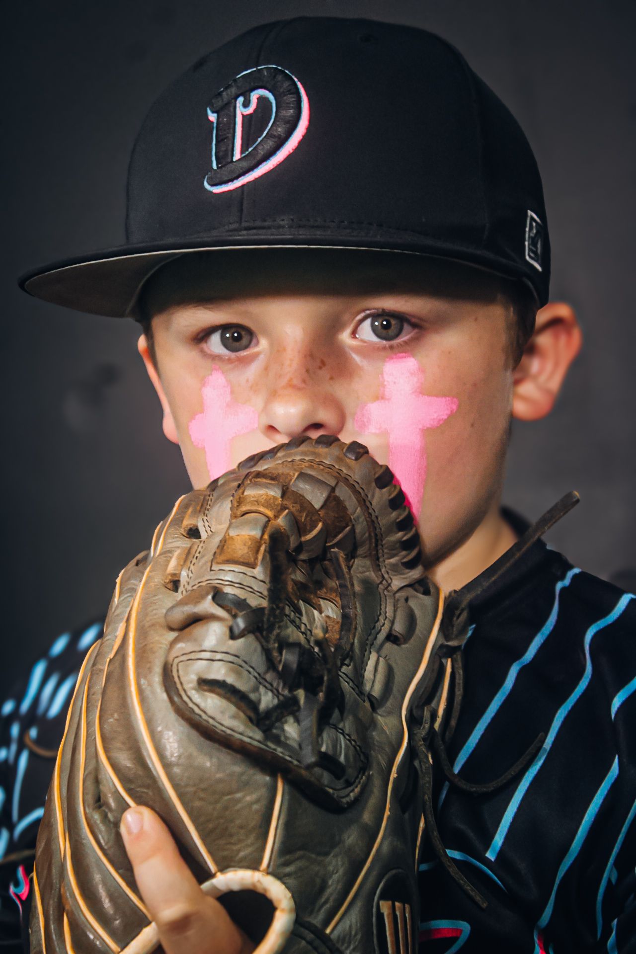 Young baseball player in a D cap holds a worn leather glove to his face with pink cross face paint.