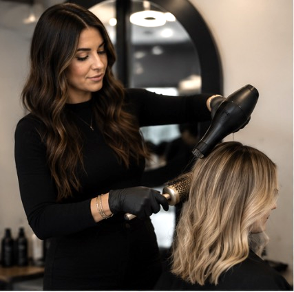 Hair stylist in black outfit blow drying client's blonde highlighted hair in modern salon.