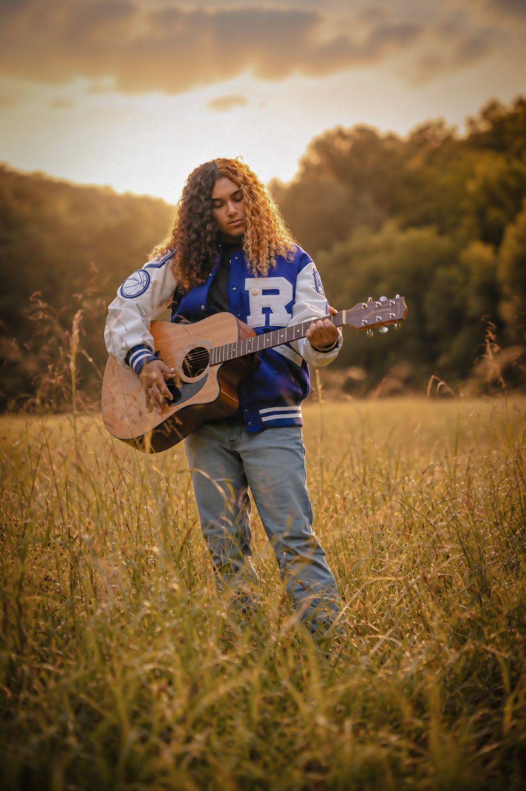 A curly-haired musician plays acoustic guitar in a golden field wearing a blue varsity jacket at sunset.