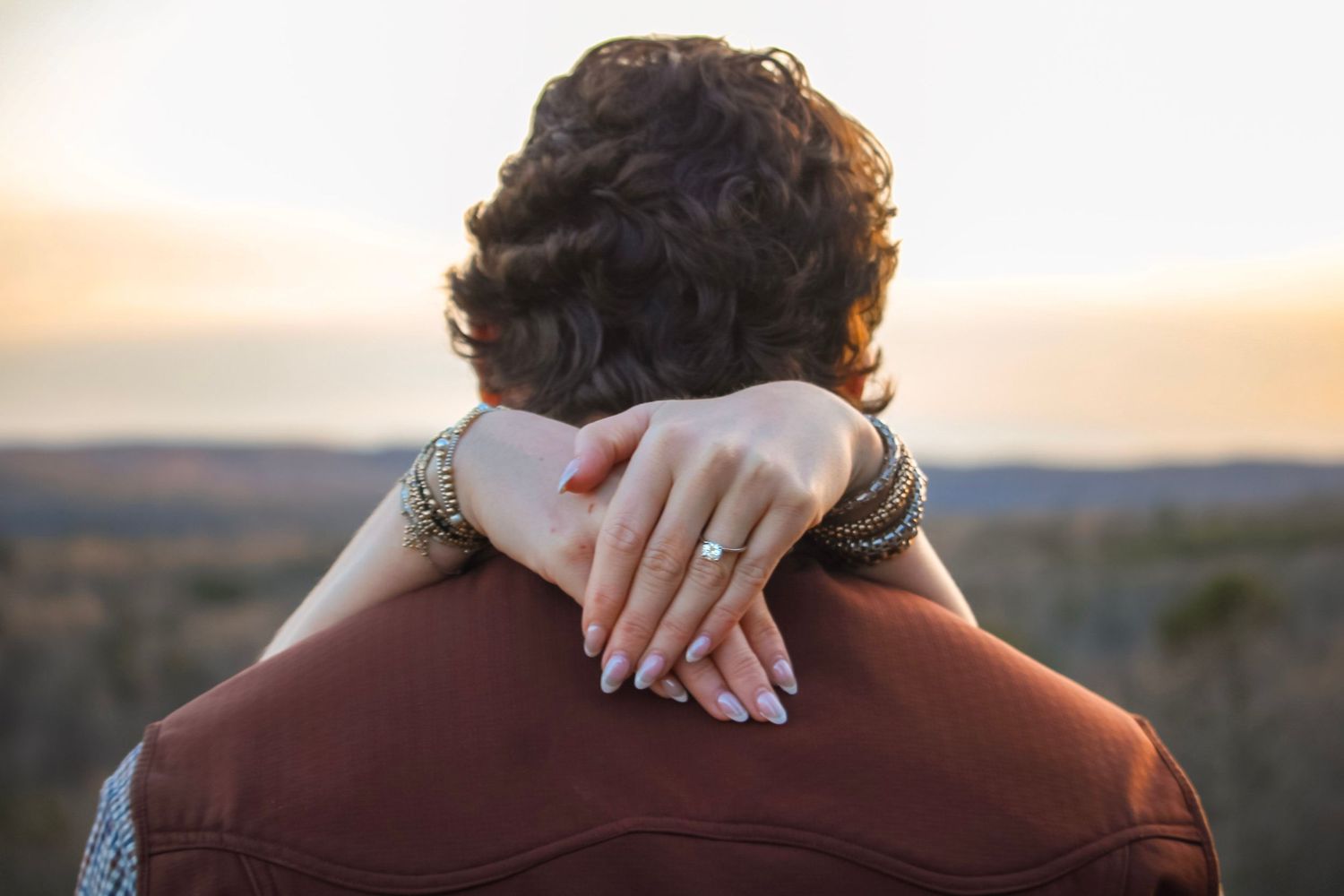 Woman resting her hand with an engagement ring on a mans shoulder at sunset.